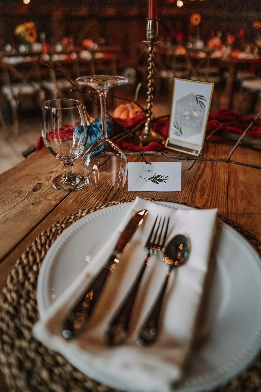 A beautifully set rustic dinner table with a white plate, silverware wrapped in a white napkin, wine glass, and water glass. The table also features a card with the name 'James Killey', a small decorative sign, candles, and natural fall-themed decor such as leaves and branches. The background shows a dimly lit, cozy dining space with wooden furniture.