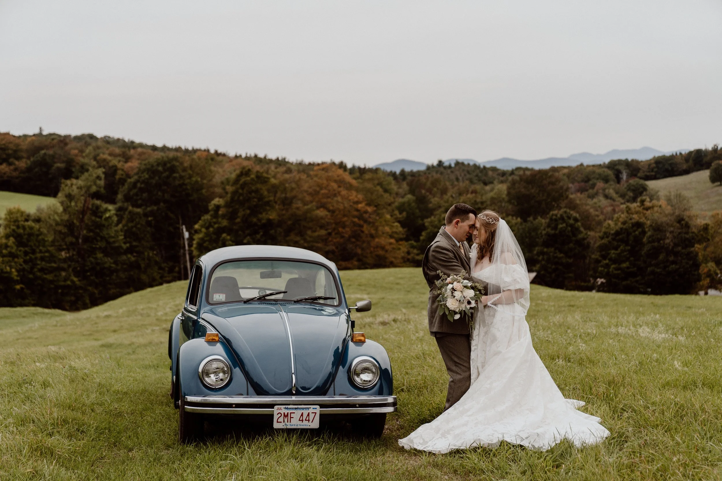 A wedding couple in wedding attire, a bride in a white gown with a veil holding a bouquet, and a groom in a brown suit, standing close together near a vintage blue car on a grassy field with trees and hills in the background.