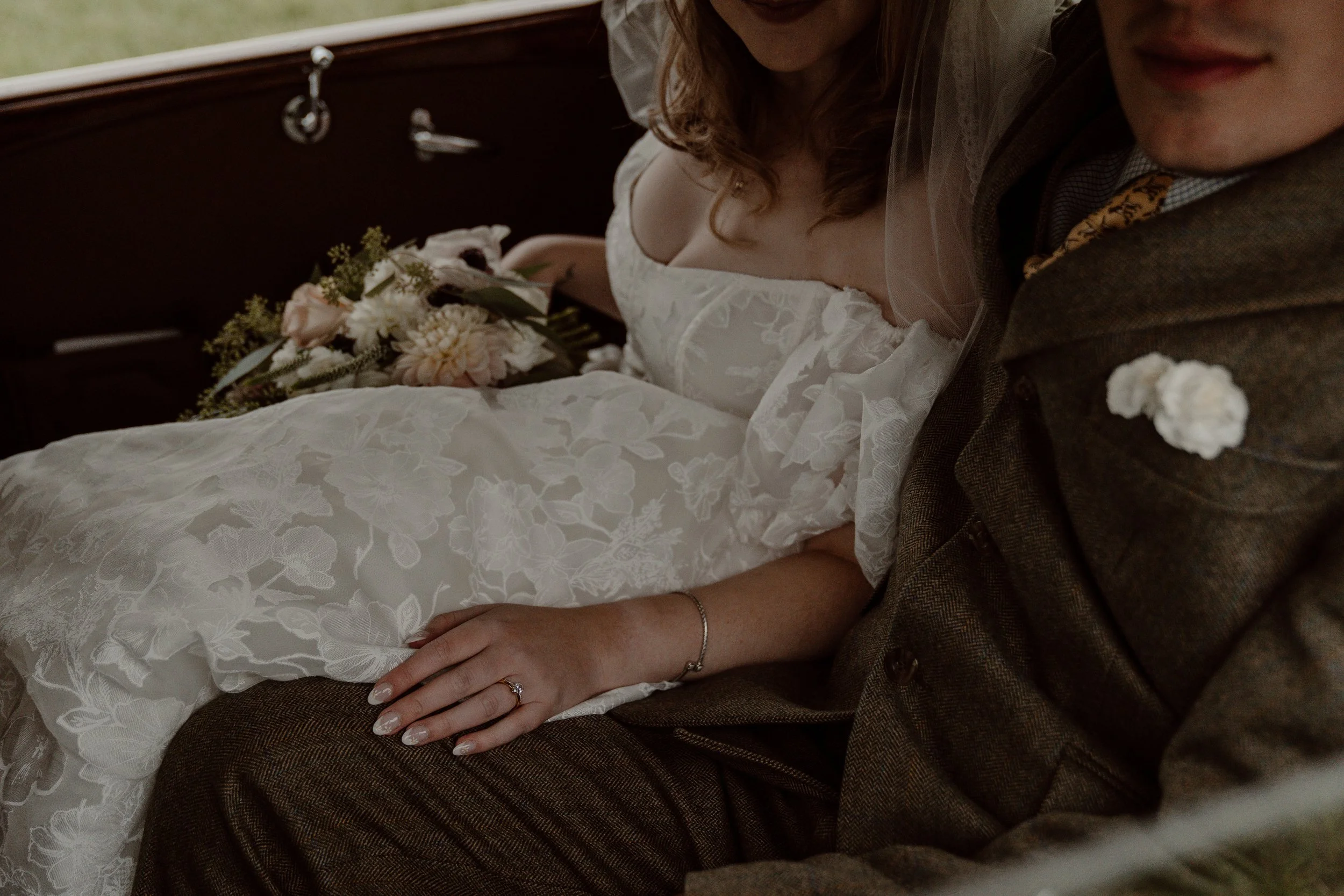 Bride and groom sitting closely, with the bride's hand resting on the groom's lap, bride in a white wedding gown holding a bouquet, groom in a brown suit, wedding setting.