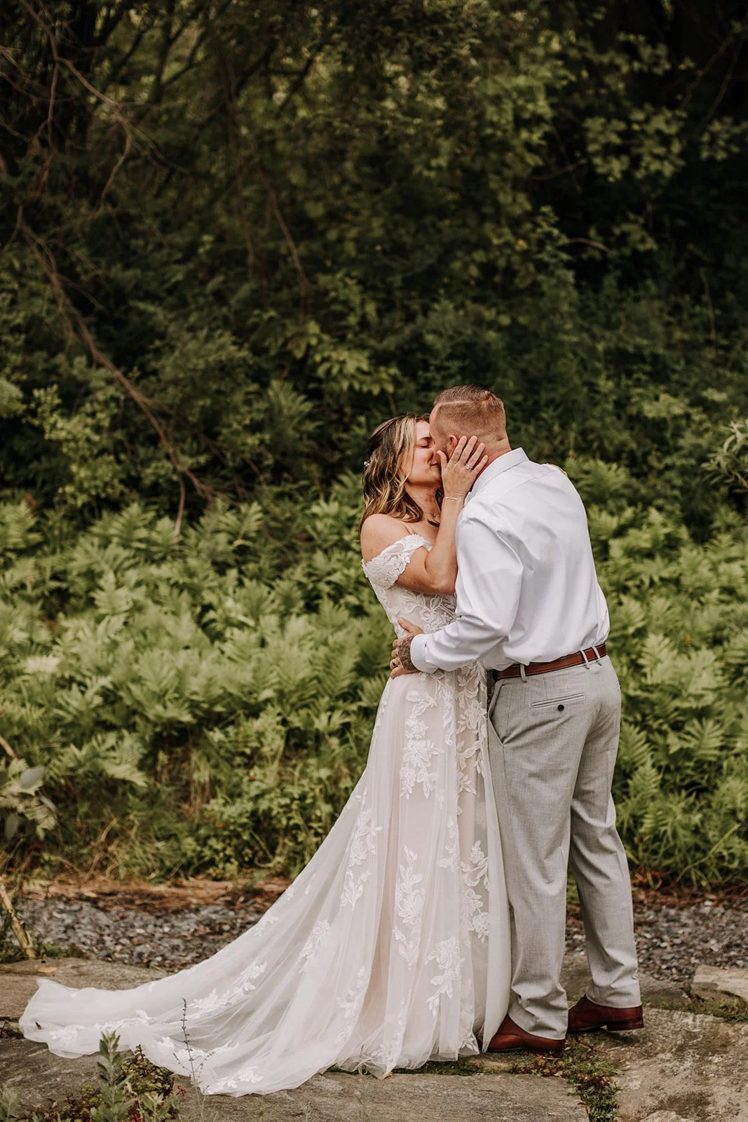 A bride and groom embrace and kiss outdoors, surrounded by lush green foliage in a natural setting.