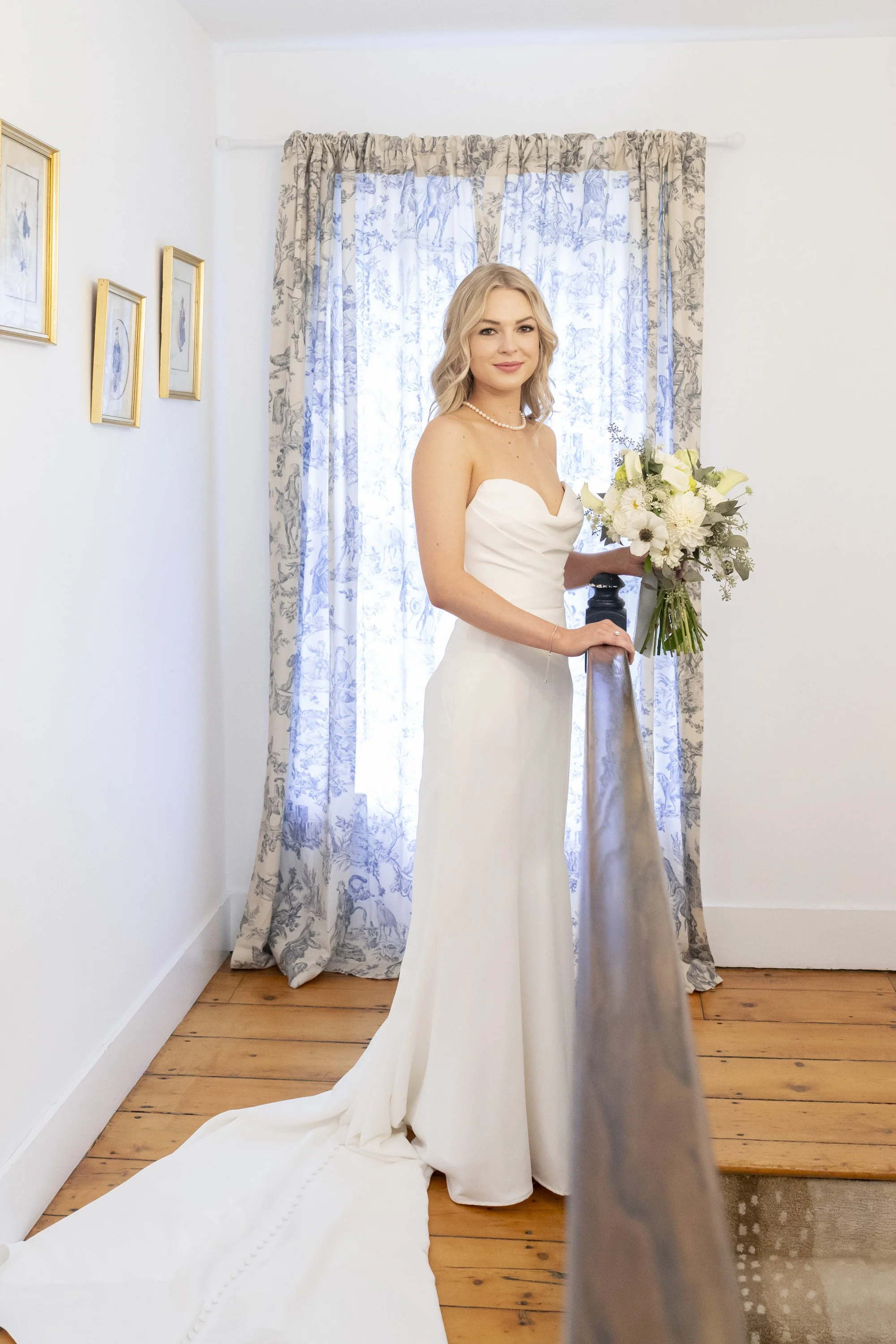 A bride in a white wedding dress holding a bouquet of white flowers standing in a room with wooden floors and floral curtains.