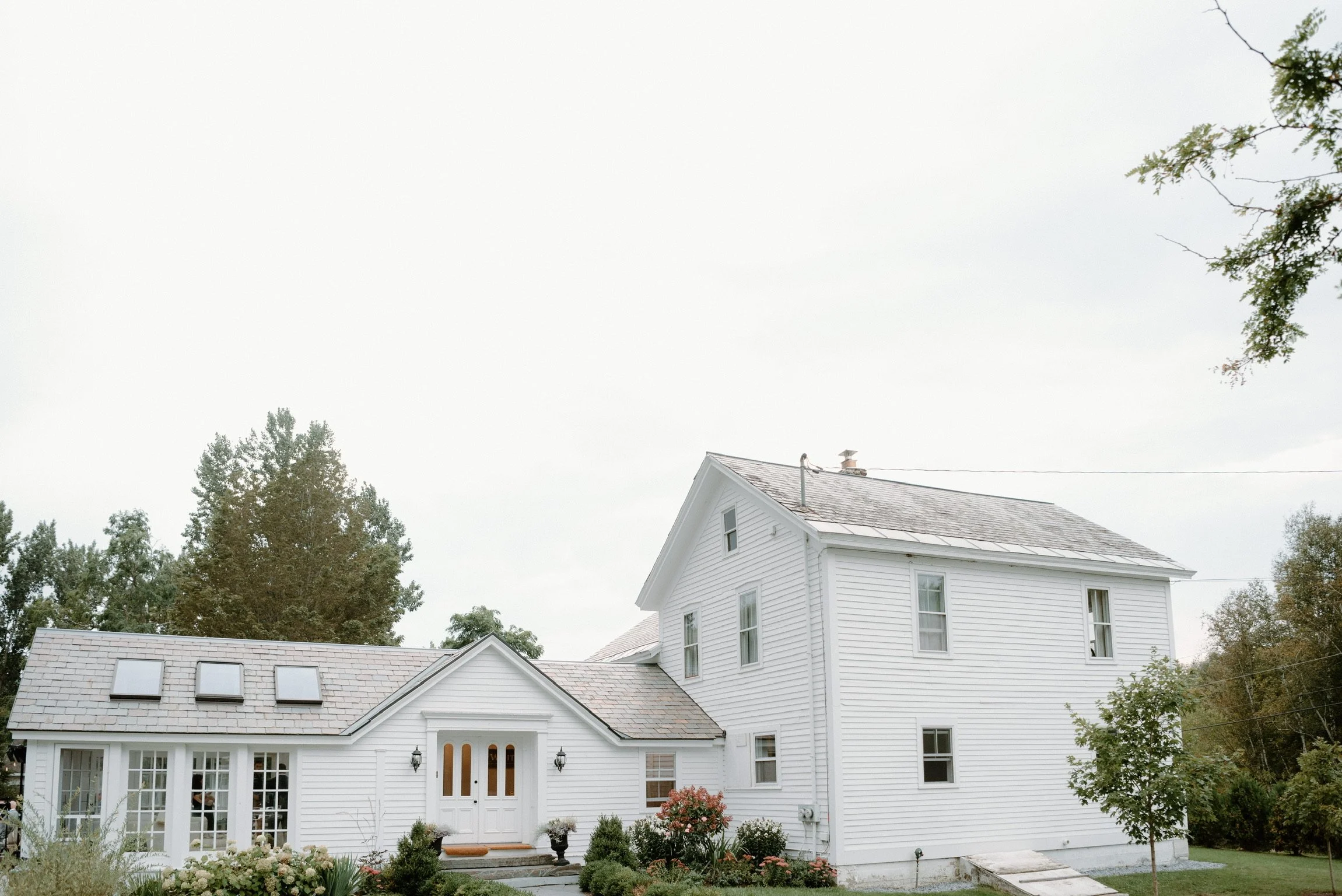 White two-story house with a gabled roof, front door, multiple windows, garden, and trees surrounded by greenery.