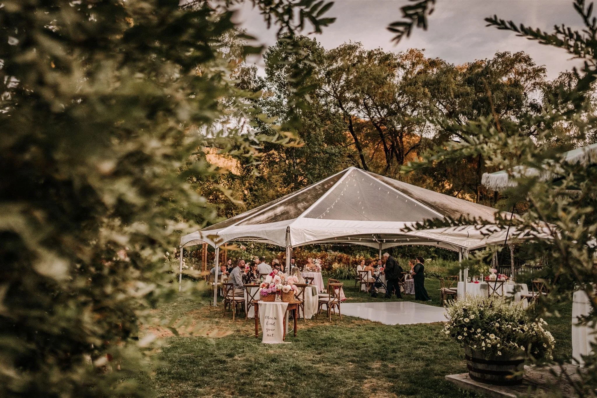 A wedding reception outdoor under a white tent, decorated with floral arrangements and string lights, with guests seated at tables. The scene is surrounded by trees and greenery, with a dance floor in the foreground.