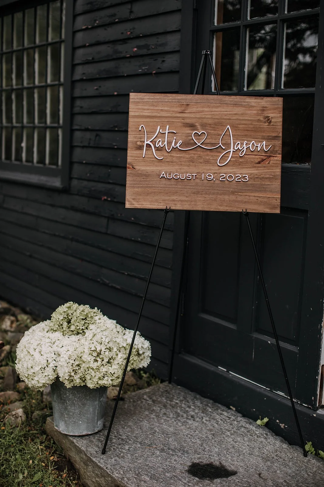 A wooden sign with white wire lettering that reads 'Katie ♥ Jason' and 'August 19, 2023' is positioned on a black easel outside a dark-colored building. A pot of white flowers sits on a stone step nearby.