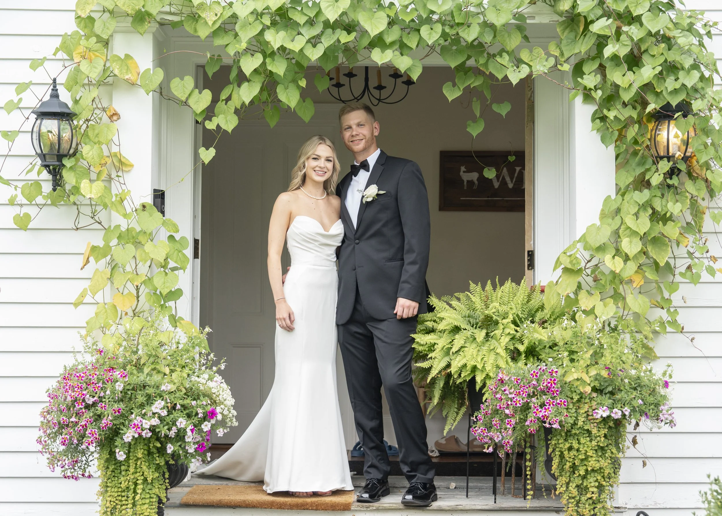 Bridal couple standing on porch with plants and greenery in the background, bride in white gown and groom in black tuxedo, smiling.
