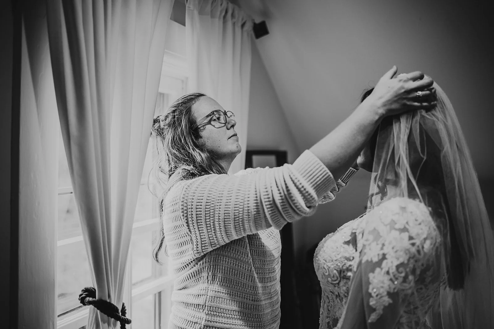 A woman adjusting a bride's veil in a room with window curtains.