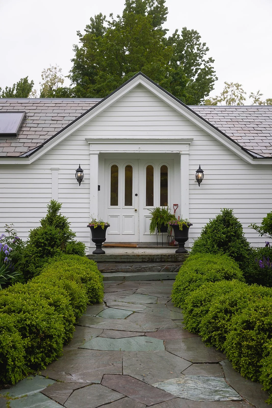 Front view of a white house with a gable roof, double front door, two black wall-mounted lantern lights, and a stone pathway lined with green shrubs leading to the entrance.