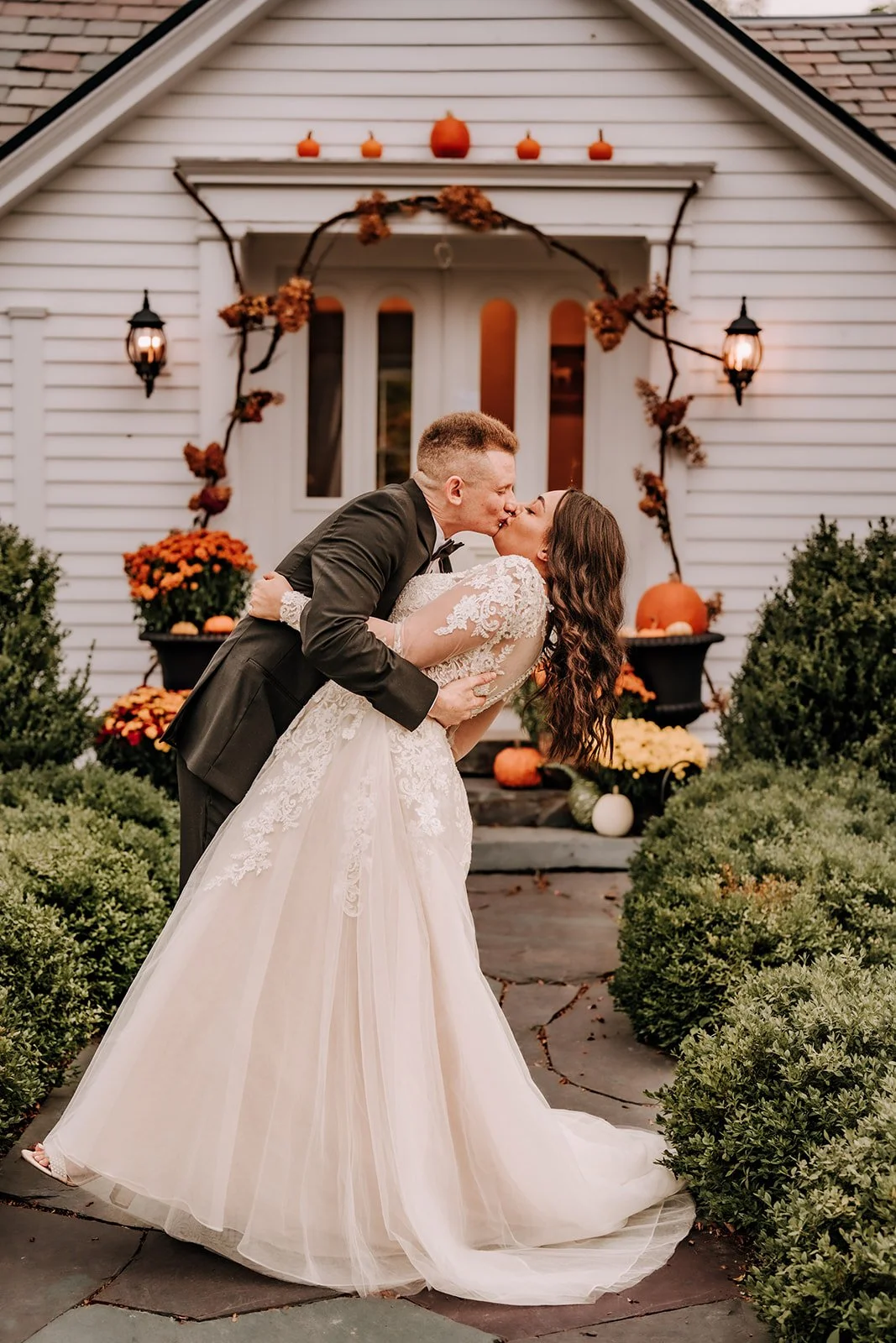 A newlywed couple sharing a kiss outside a white house decorated for fall with pumpkins and flowers, with greenery surrounding them.