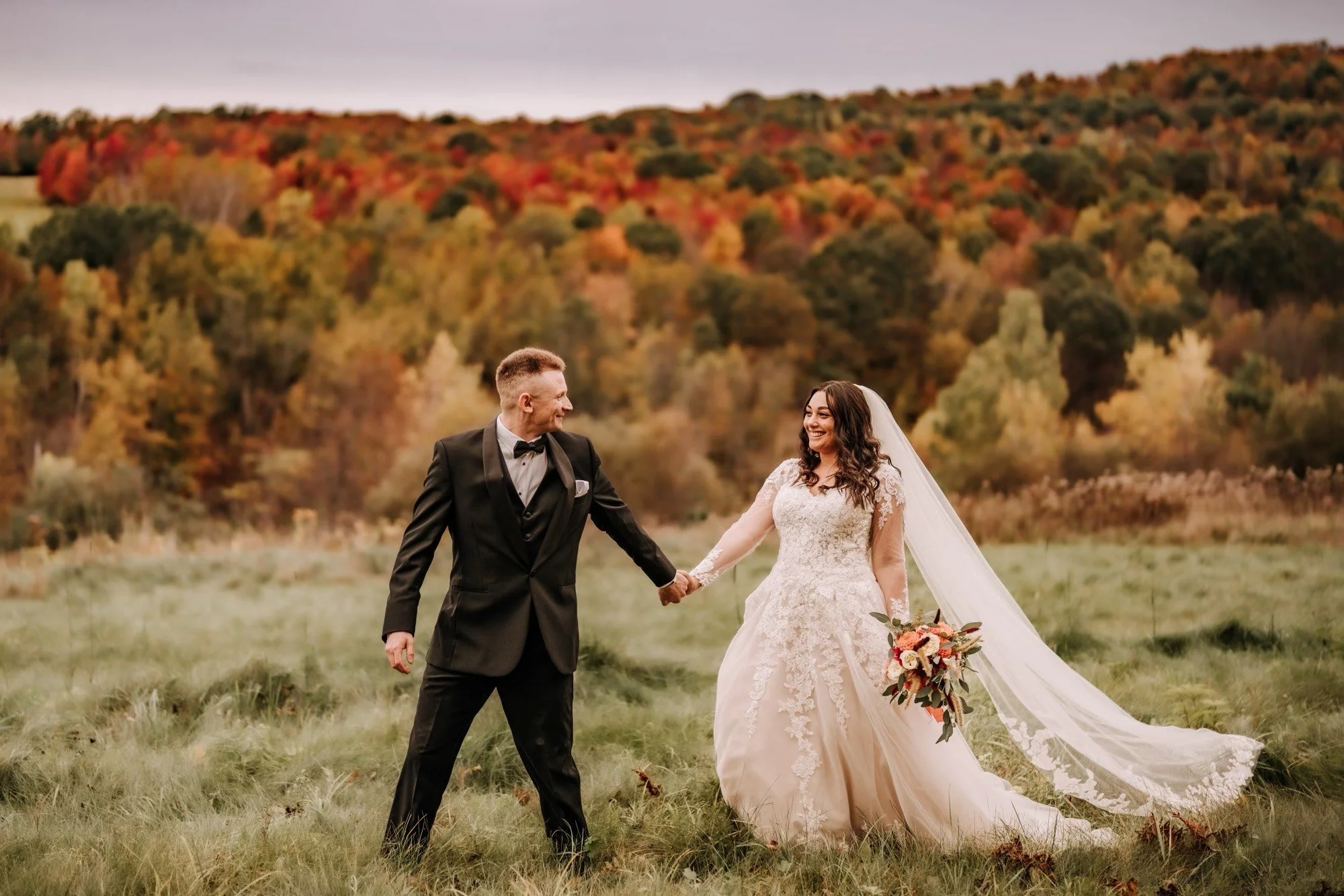A bride and groom holding hands in a field with colorful autumn trees in the background during their wedding photoshoot.