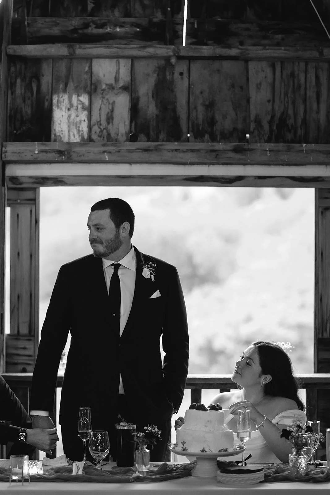 A black and white photo of a man in a tuxedo standing next to a woman sitting at a table with a wedding cake, during a wedding reception.