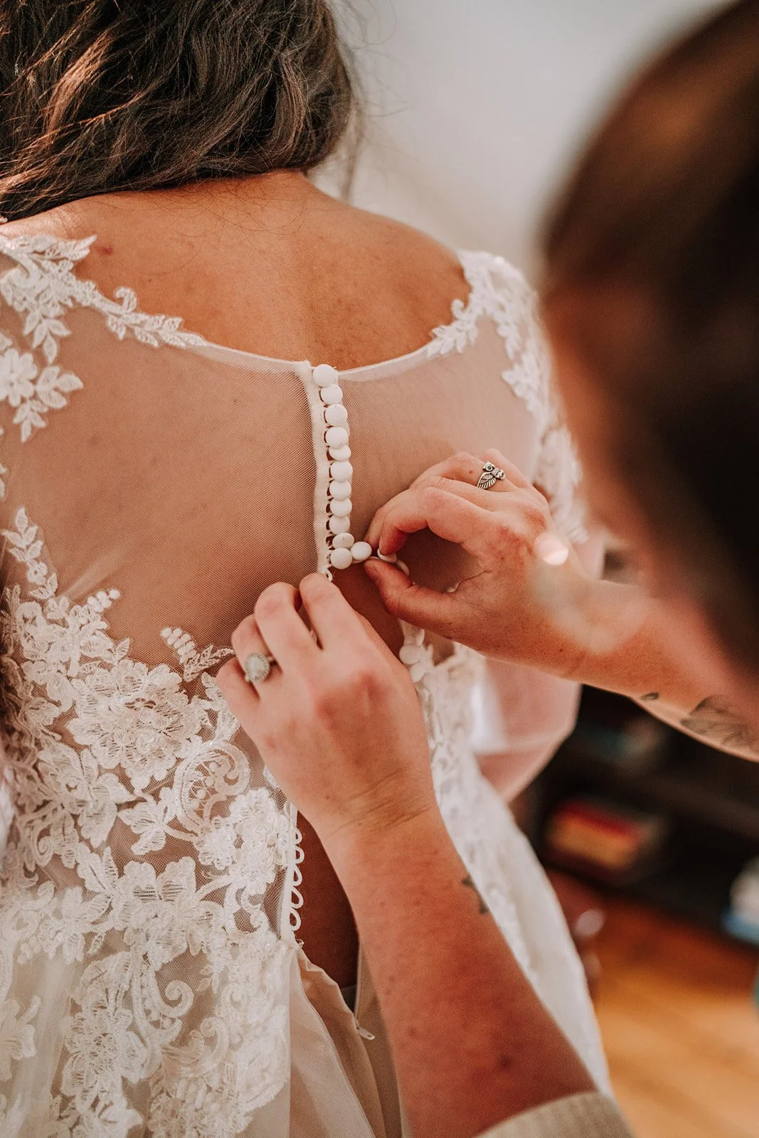 A woman in a white lace wedding dress is being buttoned up at the back by another person.
