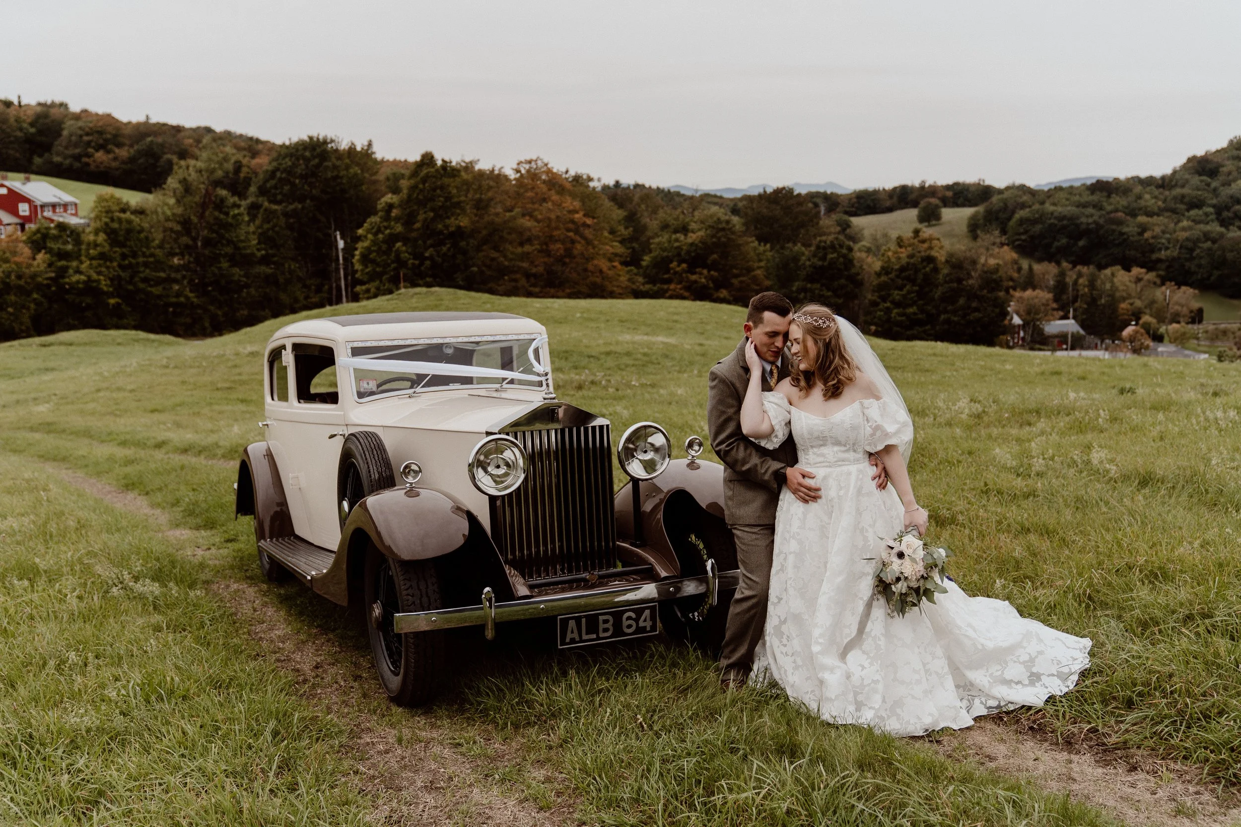 A bride and groom embrace each other beside a vintage white and black car on a grassy field, with rolling hills and trees in the background, during their wedding photoshoot.