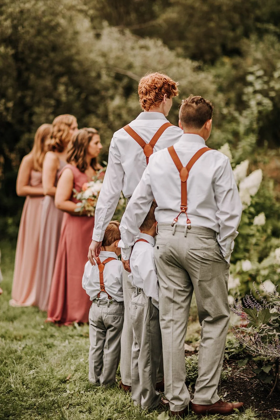 A group of people, including children and women, standing outdoors on grass, possibly waiting for a ceremony to start, with trees and white flowers in the background.