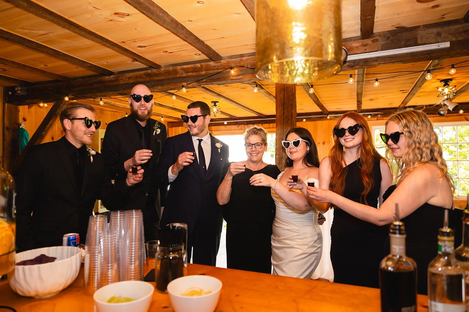 Group of seven people celebrating at a wedding reception, wearing sunglasses and raising drinks in toast inside a rustic wooden venue with string lights overhead.