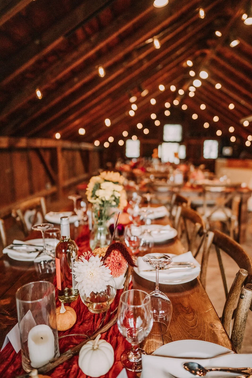 Decorated rustic dining table in a barn with string lights, floral centerpieces, pumpkins, wine glasses, and white plates.