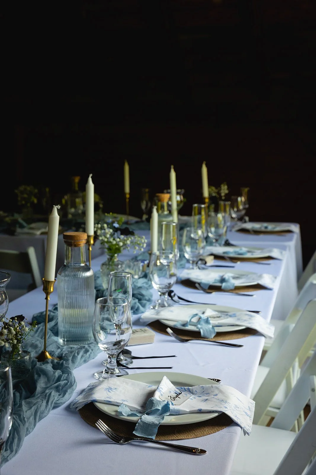 A long dining table set for a formal event with white tablecloth, gold chargers, folded napkins, glassware, tall white candles in gold holders, and small floral arrangements with white and green flowers.