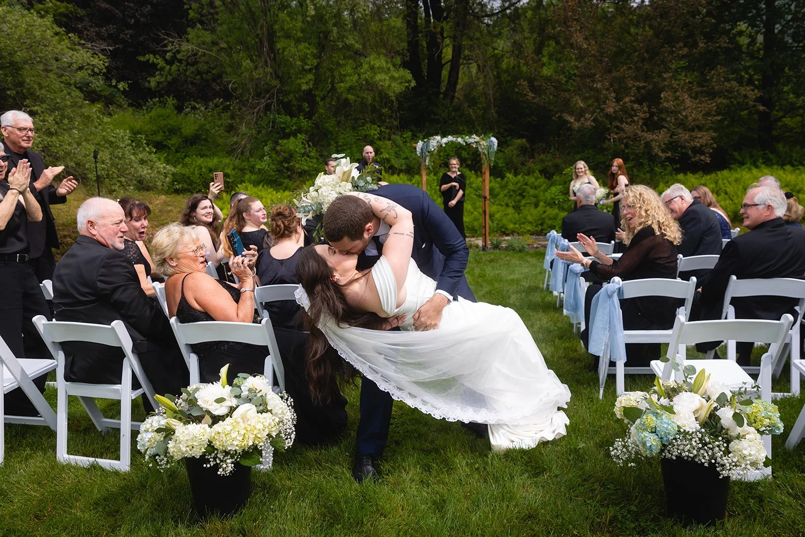 Bride and groom kissing during their outdoor wedding ceremony, with guests seated on white chairs around them and joyful expressions on their faces, set in a lush green garden.