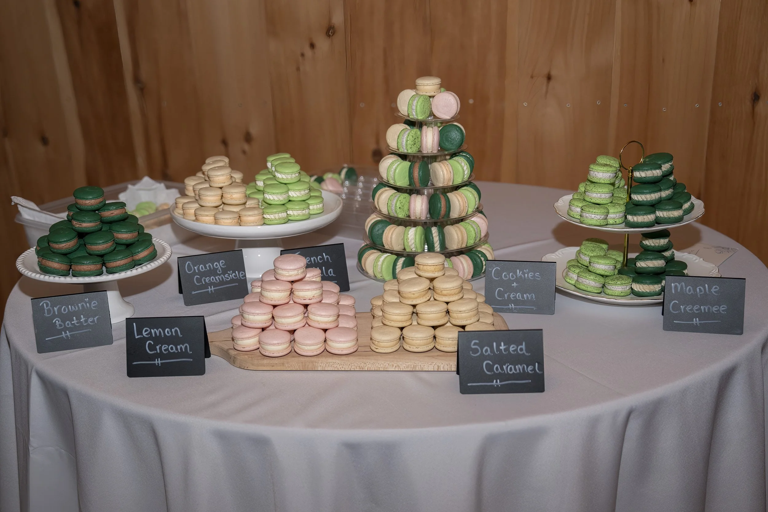 A table with a white tablecloth displaying various macarons on multiple plates and stands, with handwritten labels indicating flavors such as brown butter, lemon cream, orange creamsicle, French vanilla, salted caramel, cookies + cream, and maple creemee, against a wooden wall background.