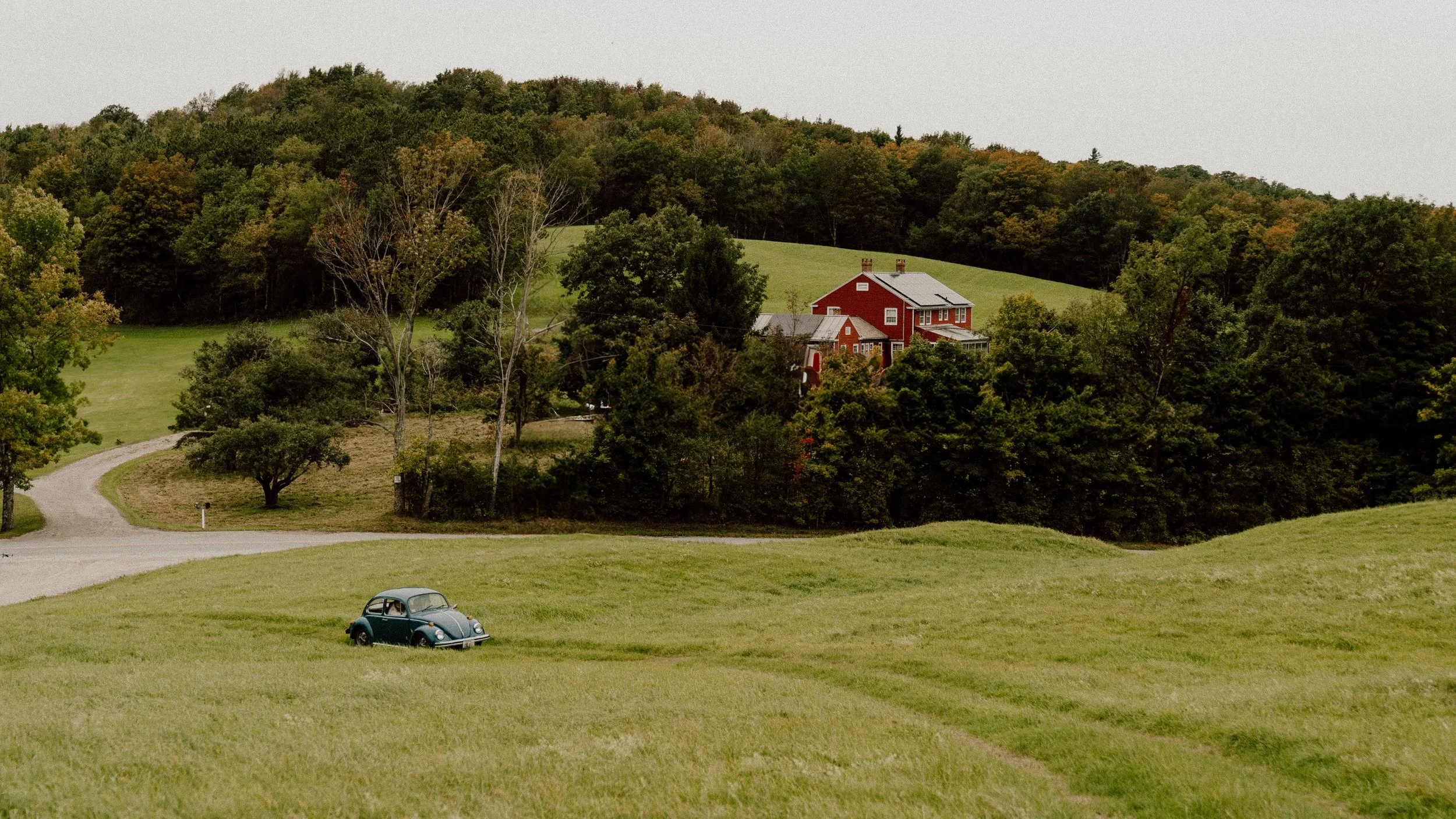 A vintage black car parked on a grassy field with a winding driveway leading to a red house on a hill surrounded by trees and rolling green landscape.