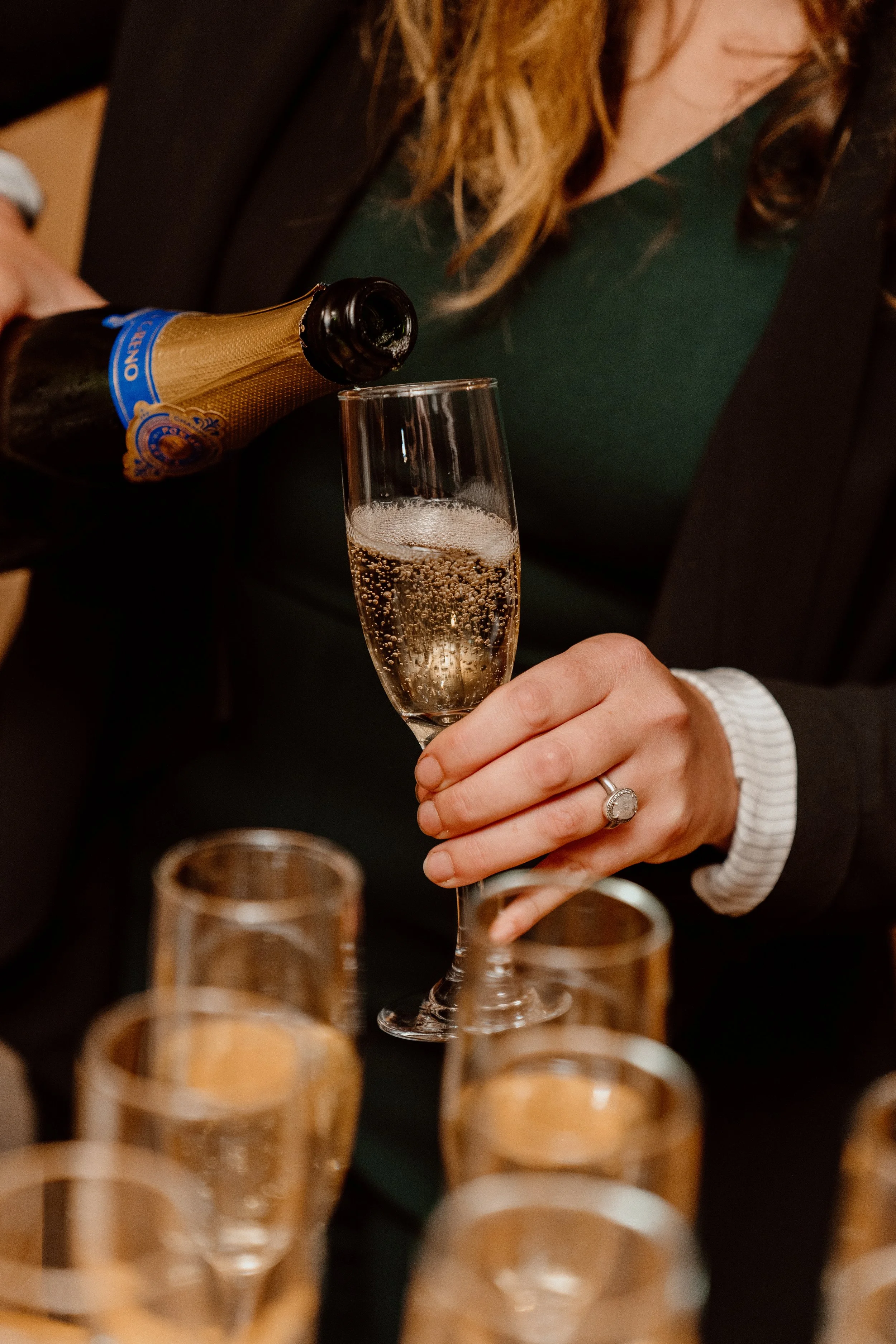 A person in a black blazer and green top is pouring champagne into a flute glass, with multiple other glasses on the table.