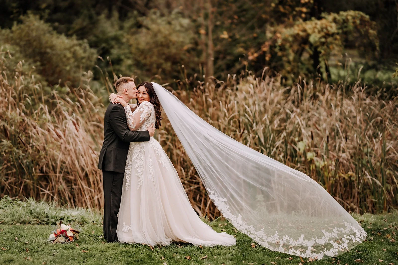 A bride and groom embrace outdoors on their wedding day, surrounded by tall brown grass and trees in the background. The bride wears a white lace wedding gown and long veil, while the groom is dressed in a black tuxedo. A bouquet lies on the grass nearby.