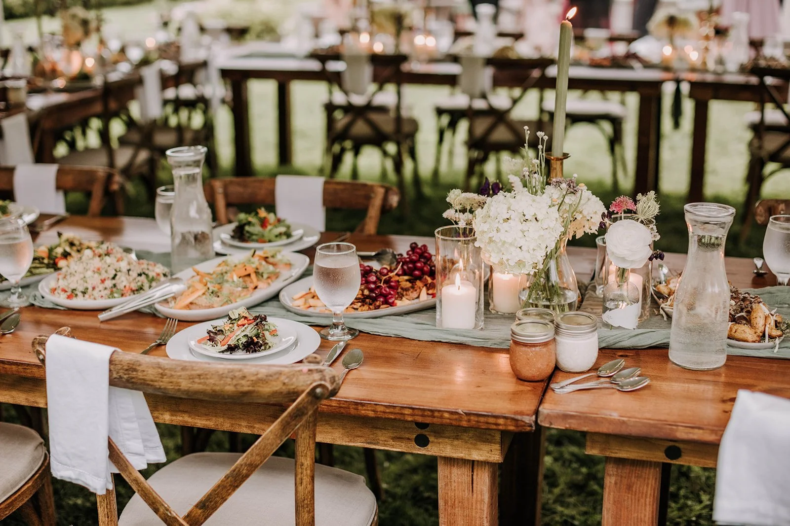 A rustic wooden outdoor banquet table set with white plates, silverware, water glasses, and floral centerpieces with candles, surrounded by wooden chairs with white napkins, and decorated for a celebration.