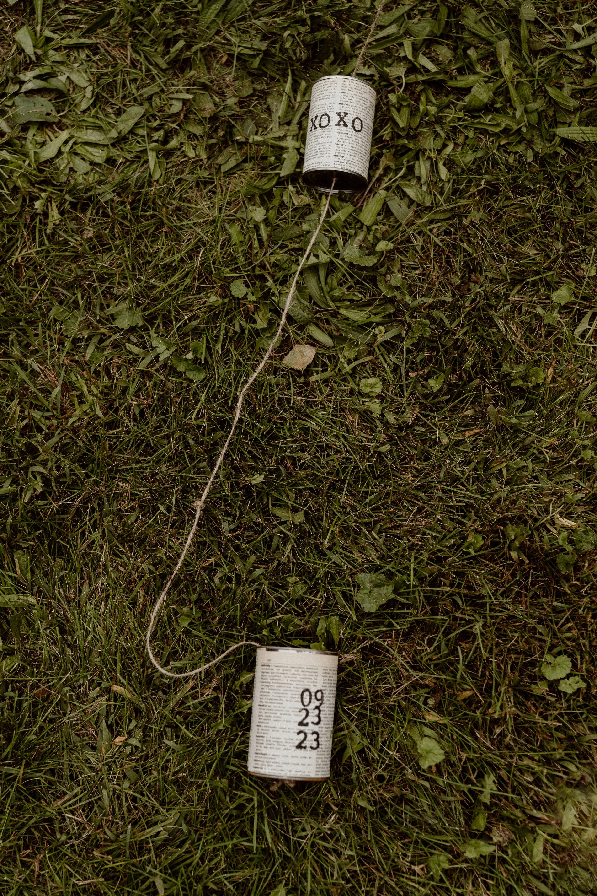 Two cans with text and numbers connected by a string lying on grass.