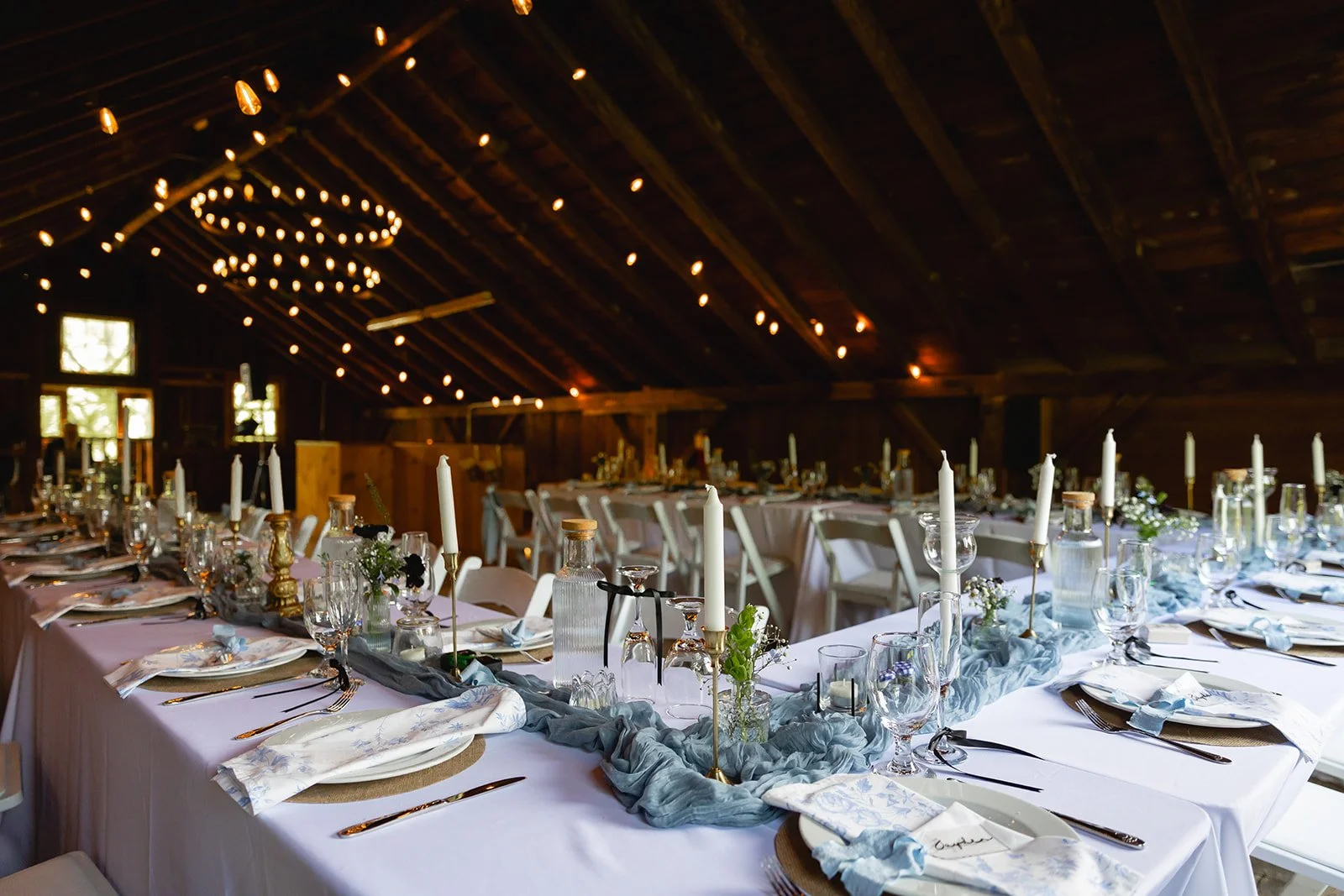 Elegant banquet table set for a celebration in a rustic barn with string lights and a chandelier.