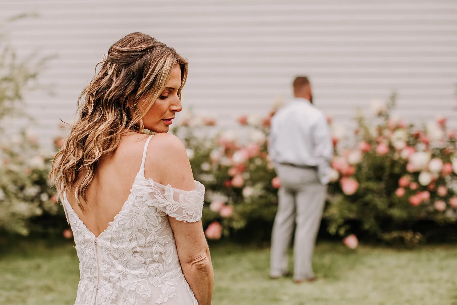 A woman in a white lace dress with cold shoulder design and spaghetti straps standing outdoors near pink roses, with a man in a white shirt and light-colored pants in the background.