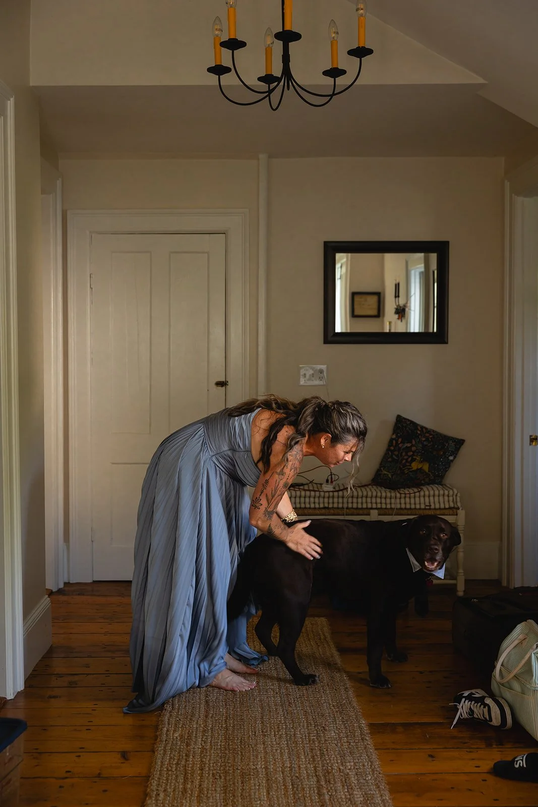 A woman with long dark hair and tattoos, wearing a long, flowing gray dress, is bending down and smiling while hugging a large black dog in a home interior with wooden floors and cream-colored walls.