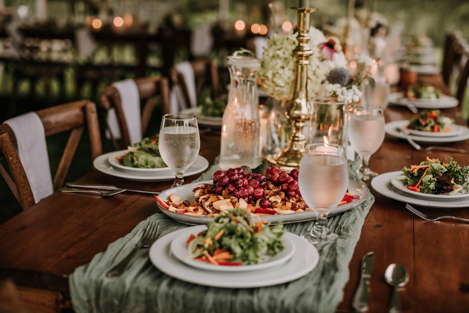 A wooden outdoor dining table set for a celebration with plates of salad, a large serving platter of assorted cheeses and grapes, water glasses, and floral centerpieces with candles.