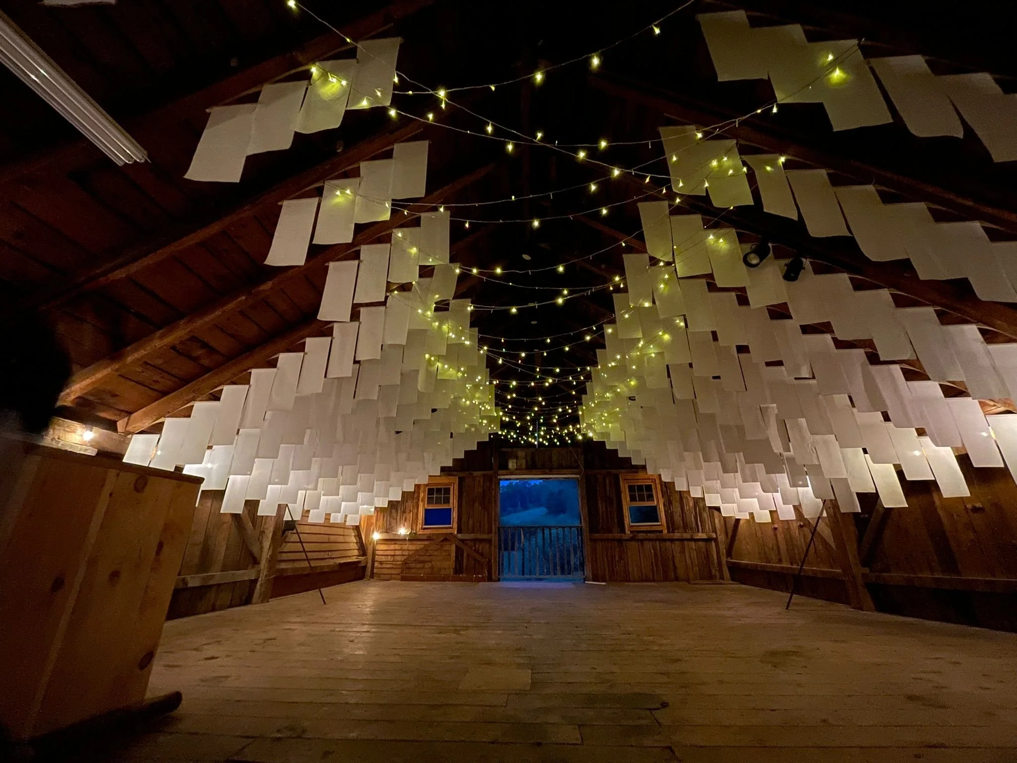 Interior of a rustic wooden barn decorated with string lights and hanging sheets of paper, with a view looking toward the open barn door at dusk.
