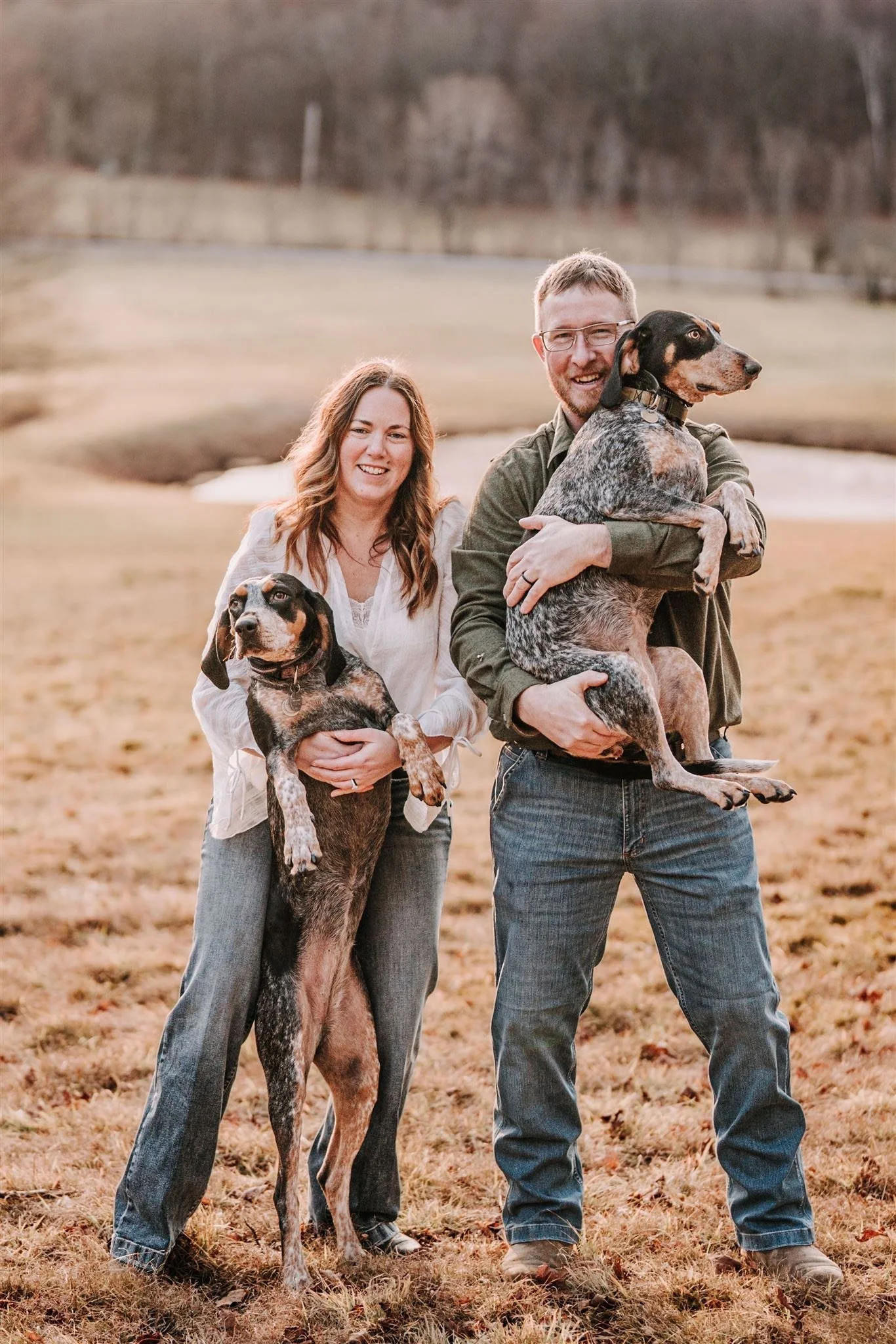 A smiling man and woman outdoors holding two dogs on a grassy field with a pond and trees in the background.