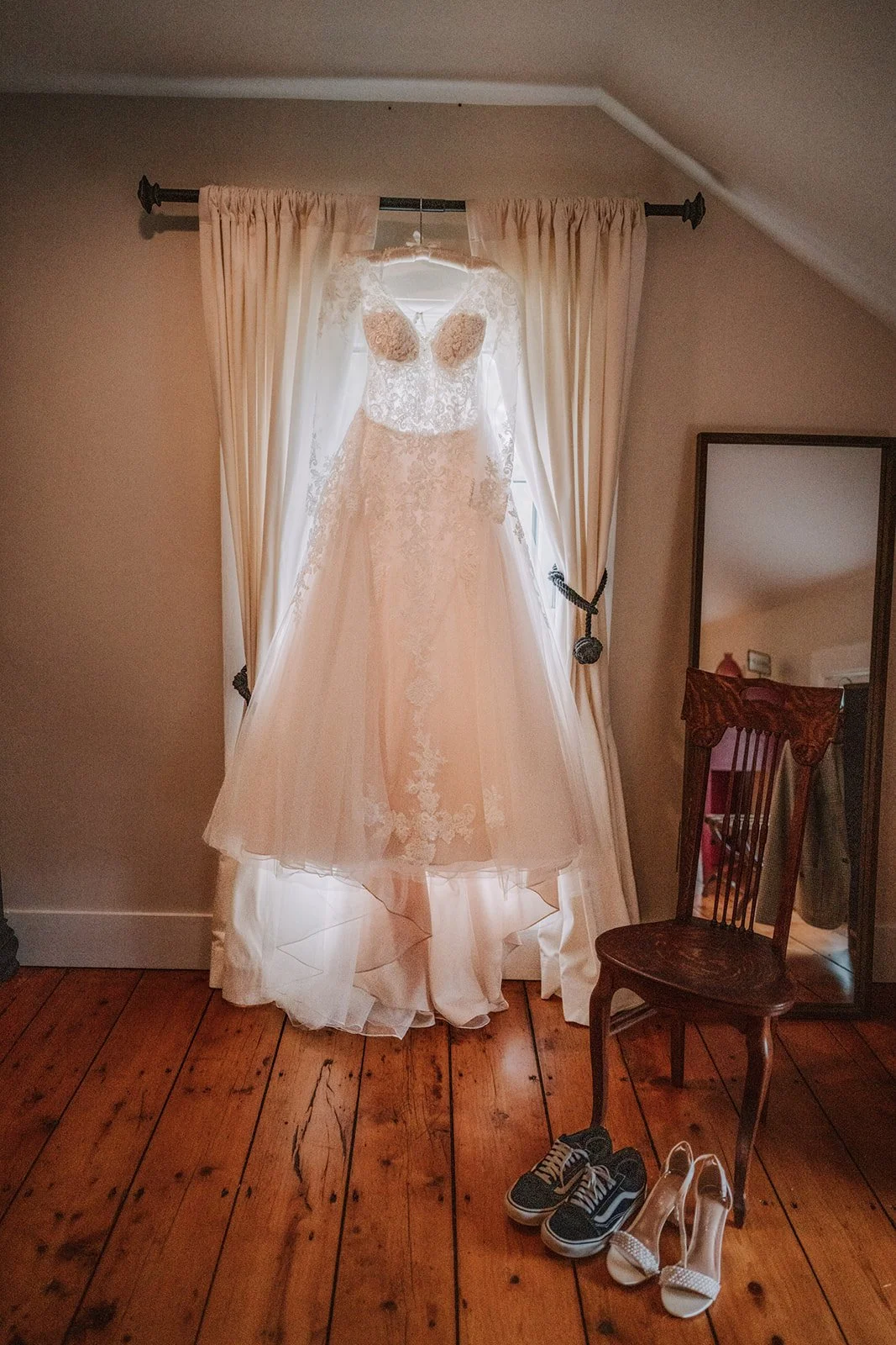 A wedding dress hanging in front of a window with cream-colored curtains, accompanied by a pair of sneakers and high heels on a wooden floor, with a chair and a mirror nearby.