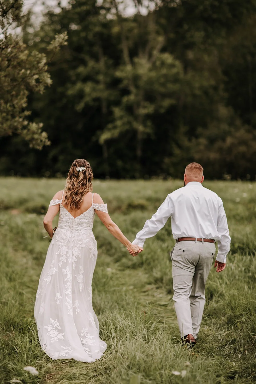 A couple, the woman in a white wedding dress and the man in a white shirt and grey pants, holding hands and walking away on a grassy field.