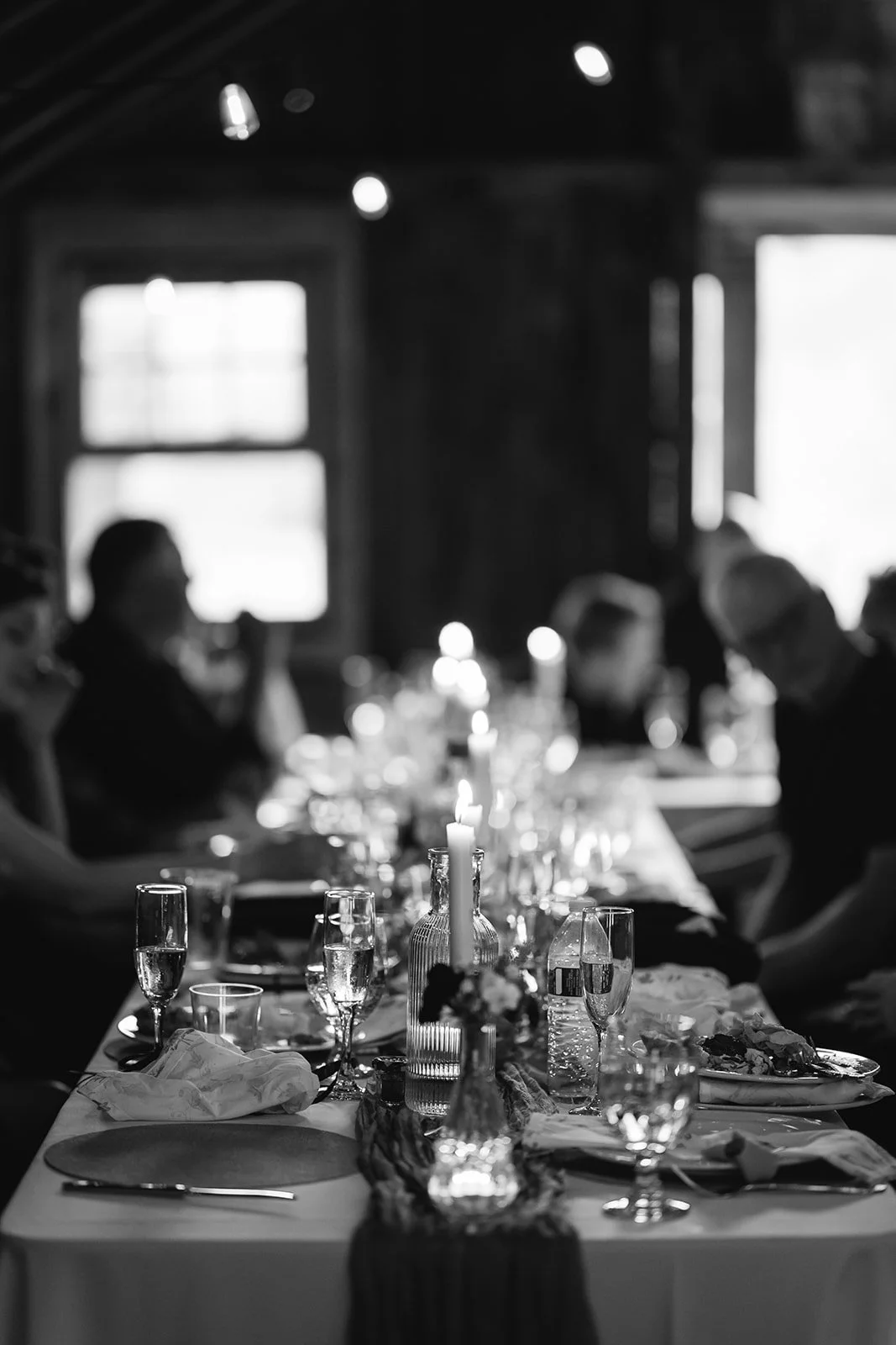 A black and white photo of a dining table set for a meal, with glasses, plates, napkins, and candles, inside a rustic room with large windows.