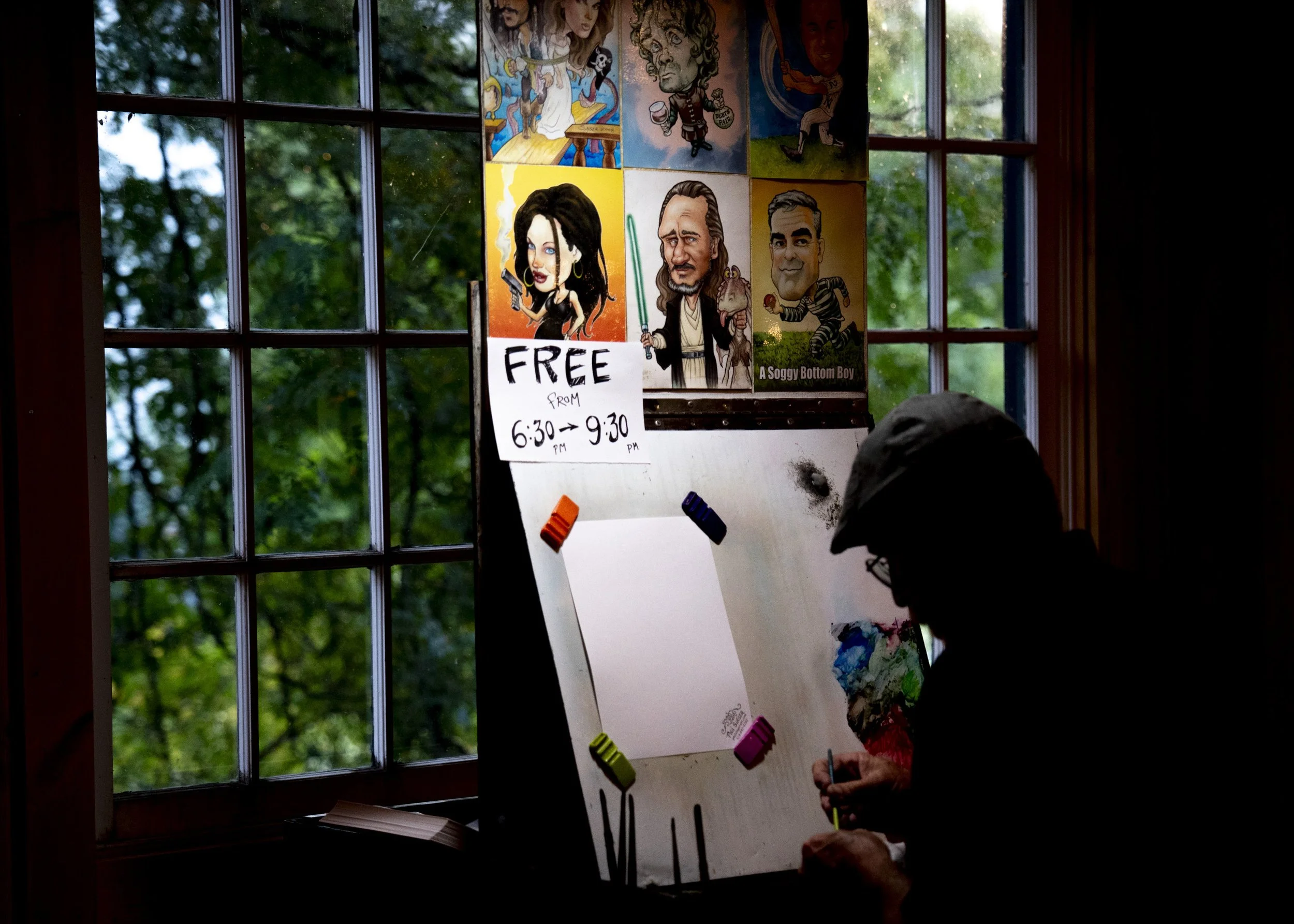 Person sketching at a whiteboard next to a window with green trees outside. The board has colorful clamped papers and caricatures of people, with a sign offering free use from 6:30 to 9:30.