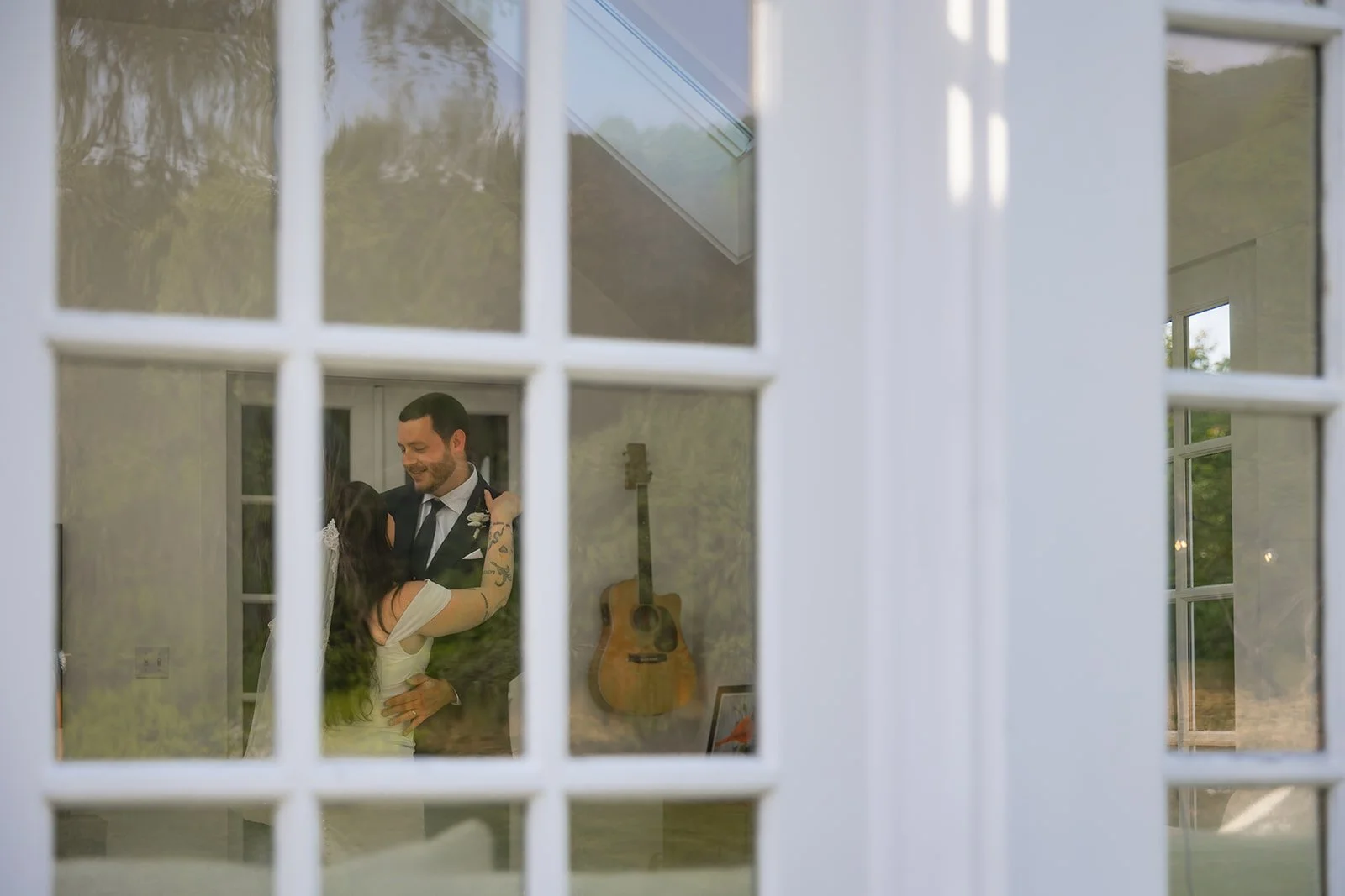 A man and woman are dancing and smiling inside a room, viewed through a glass door with a grid pattern. The room has a guitar mounted on the wall and a framed picture.