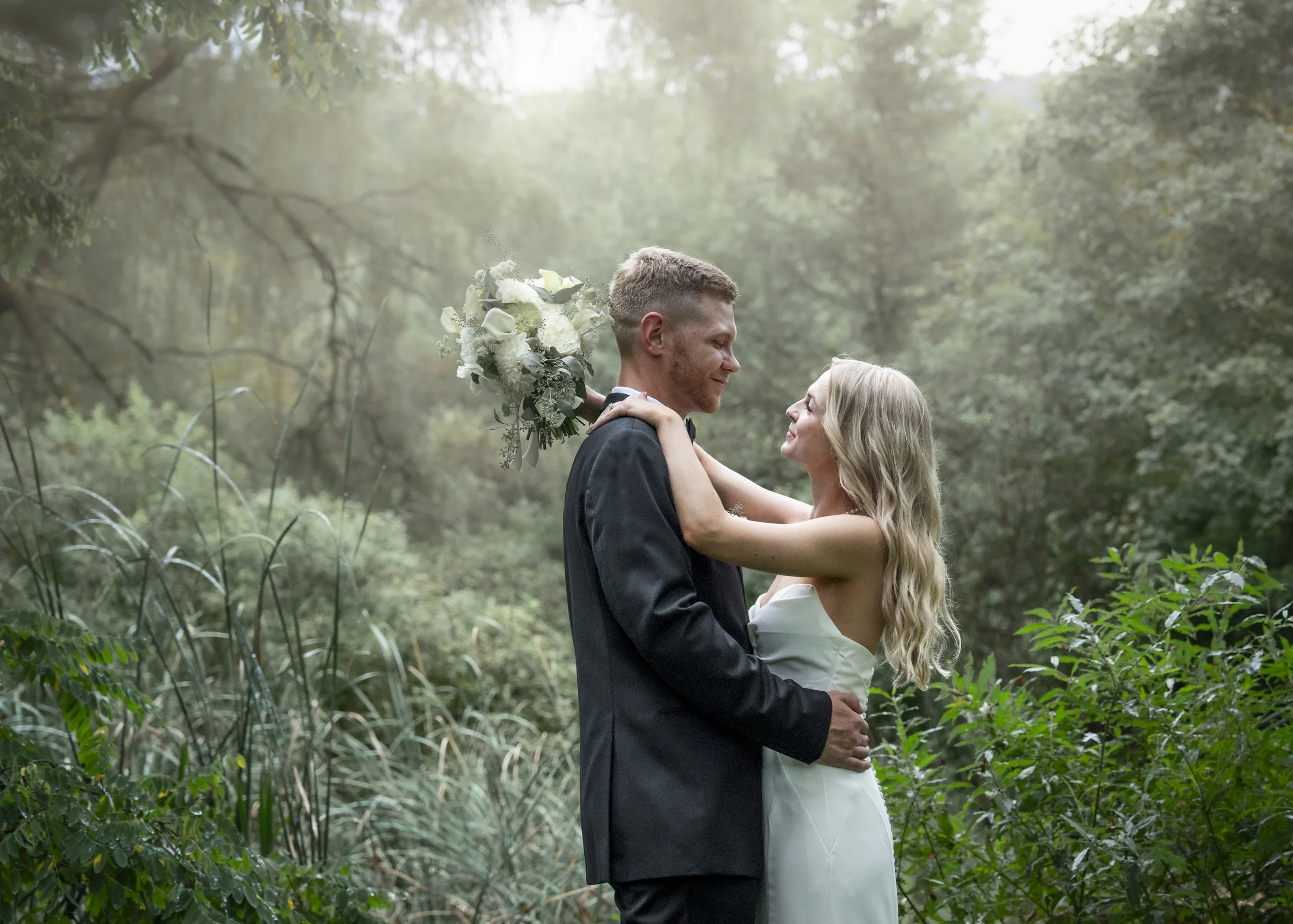 A bride and groom embrace in a forest, with the bride holding a bouquet of white flowers.