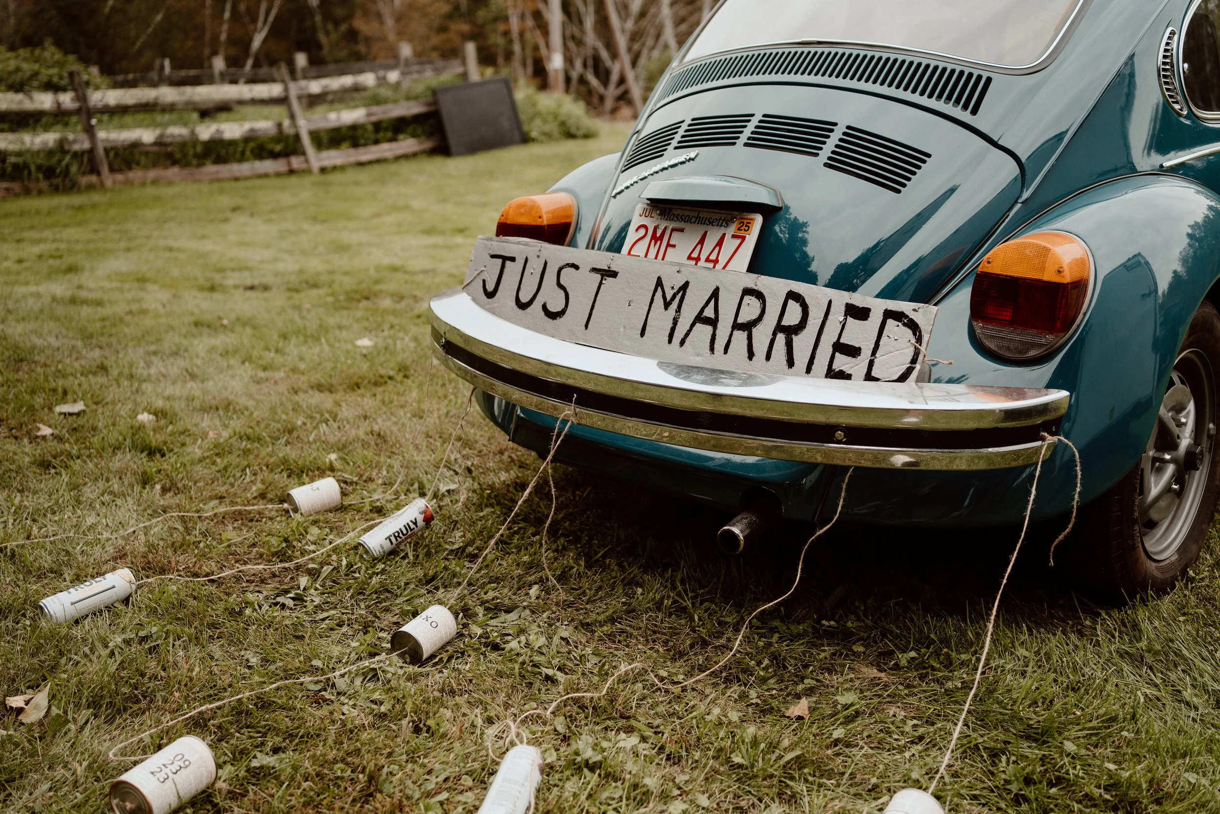 Rear view of a vintage blue Volkswagen Beetle with a "Just Married" sign on the bumper and cans tied to the bumper, seen on a grassy lawn.