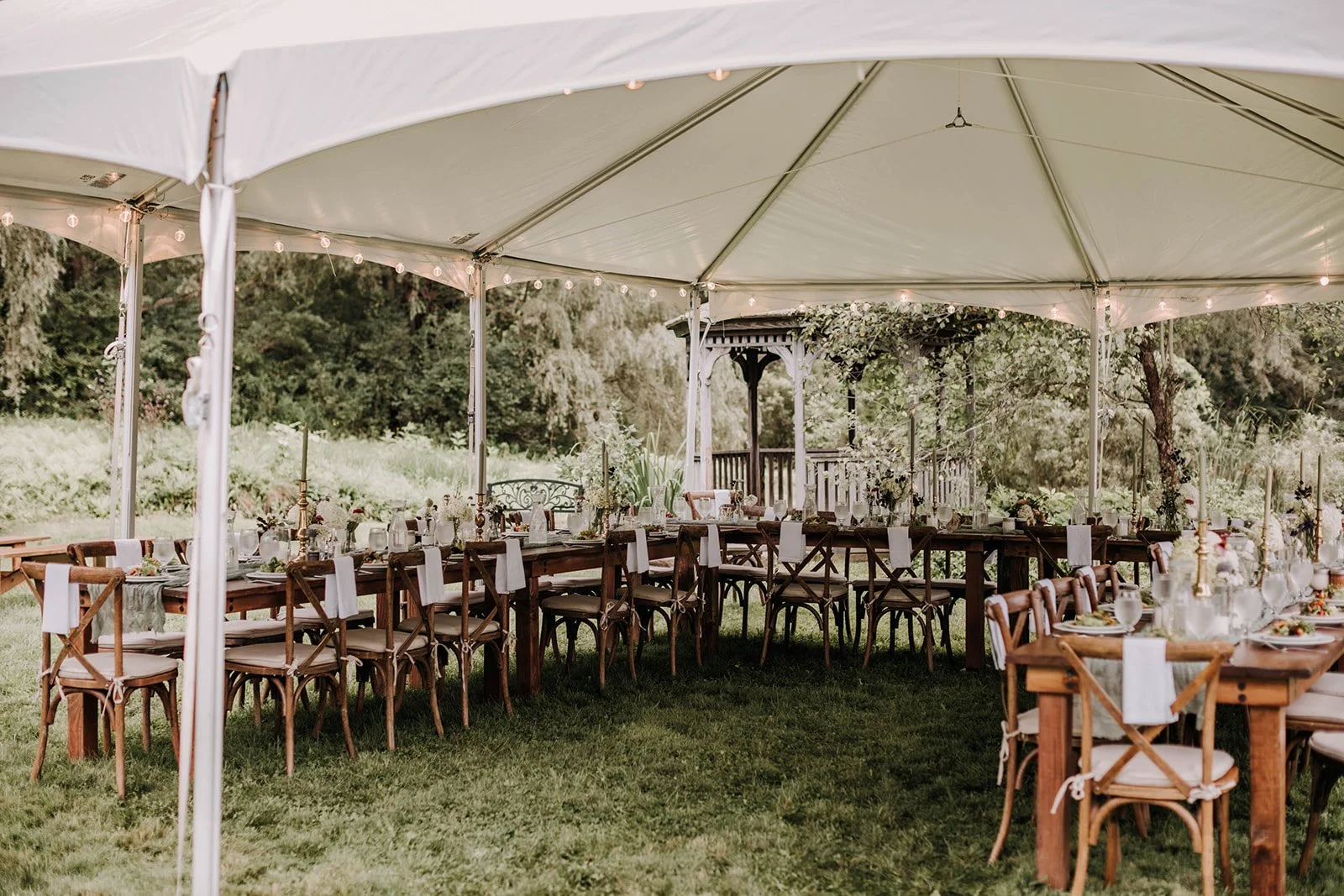 Outdoor wedding or event reception setup under a large white tent with long wooden tables, white tablecloths, and chairs, surrounded by greenery and trees.