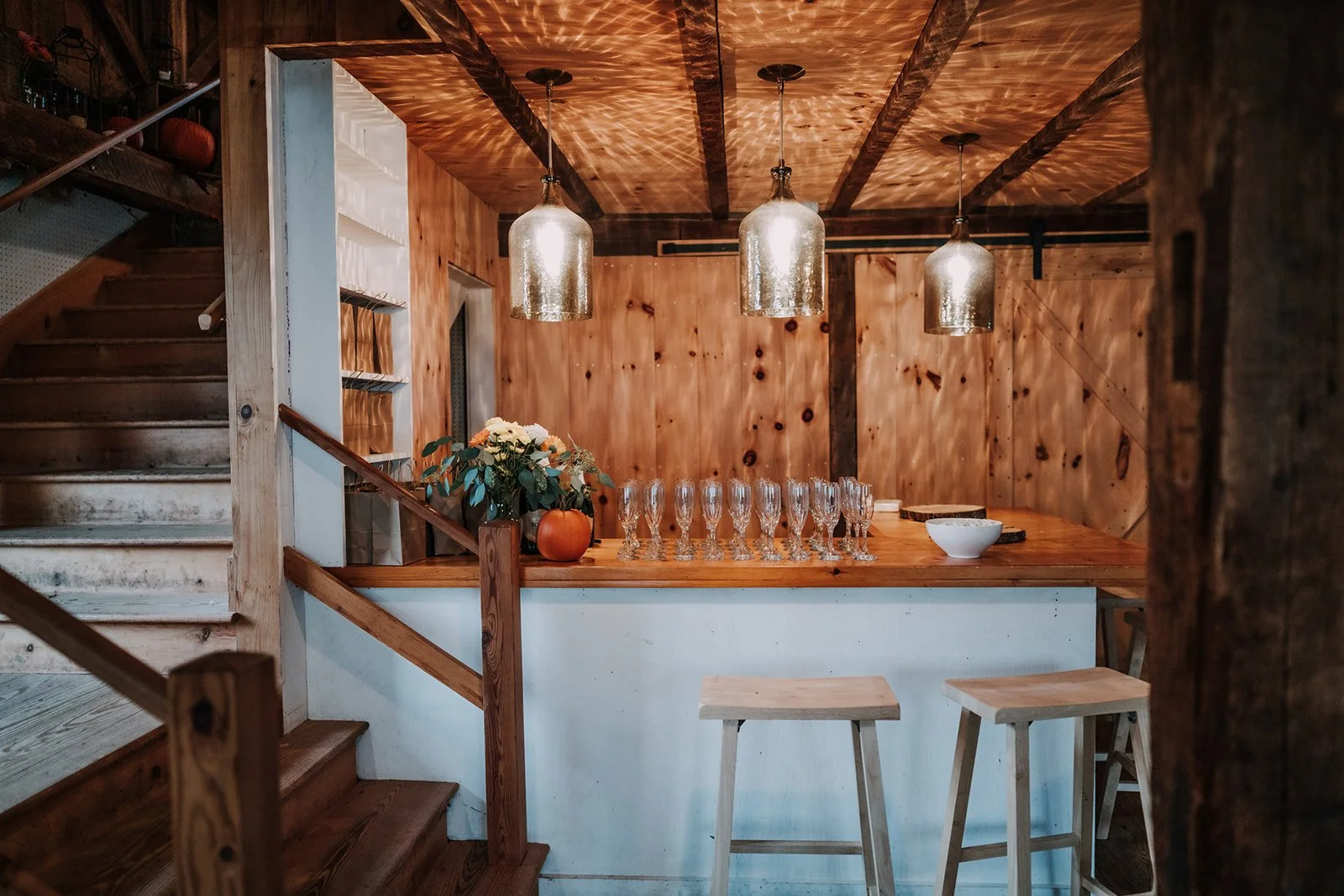 Rustic kitchen or bar area with wooden paneling, three hanging pendant lights, a counter with champagne glasses, a bowl, and flower arrangement with a pumpkin, with wooden stools and a staircase.