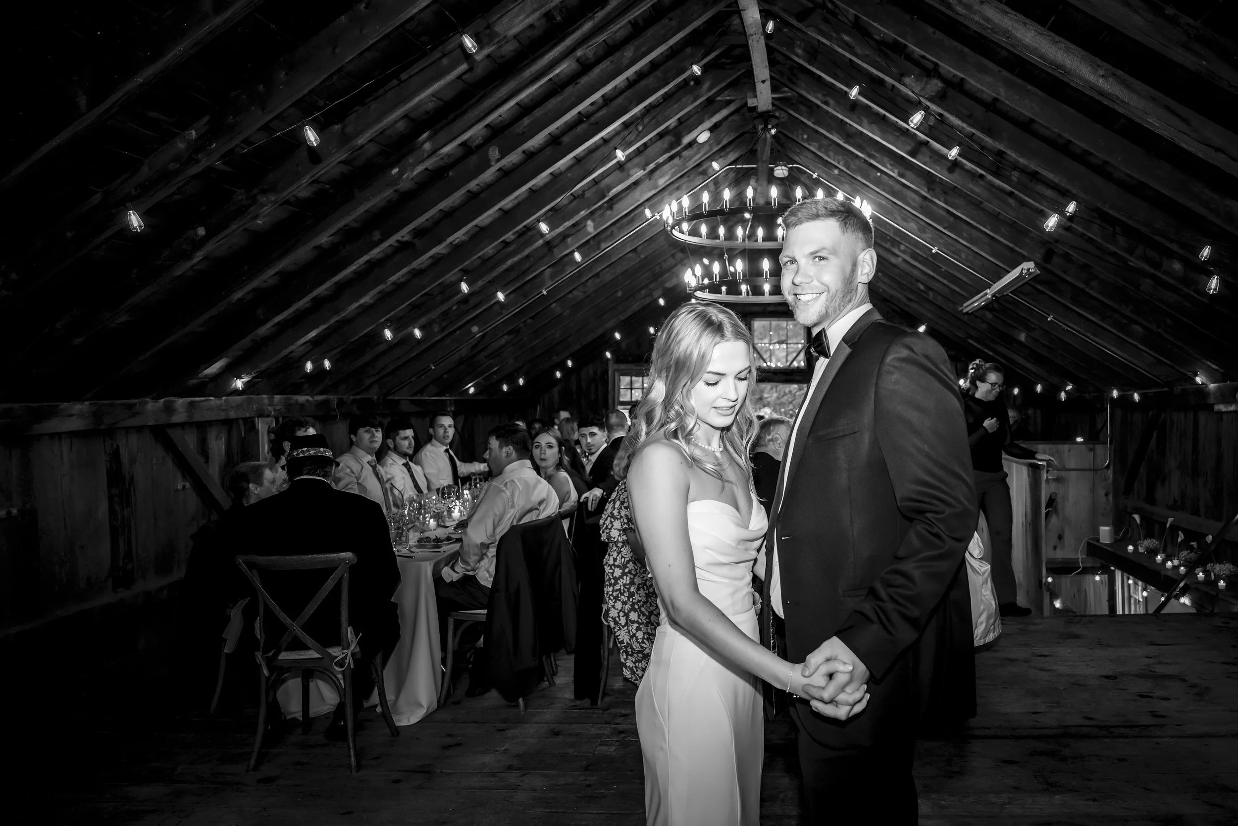 A bride and groom dance at their wedding reception in a rustic barn with wooden ceiling, chandelier, and string lights, surrounded by seated guests.