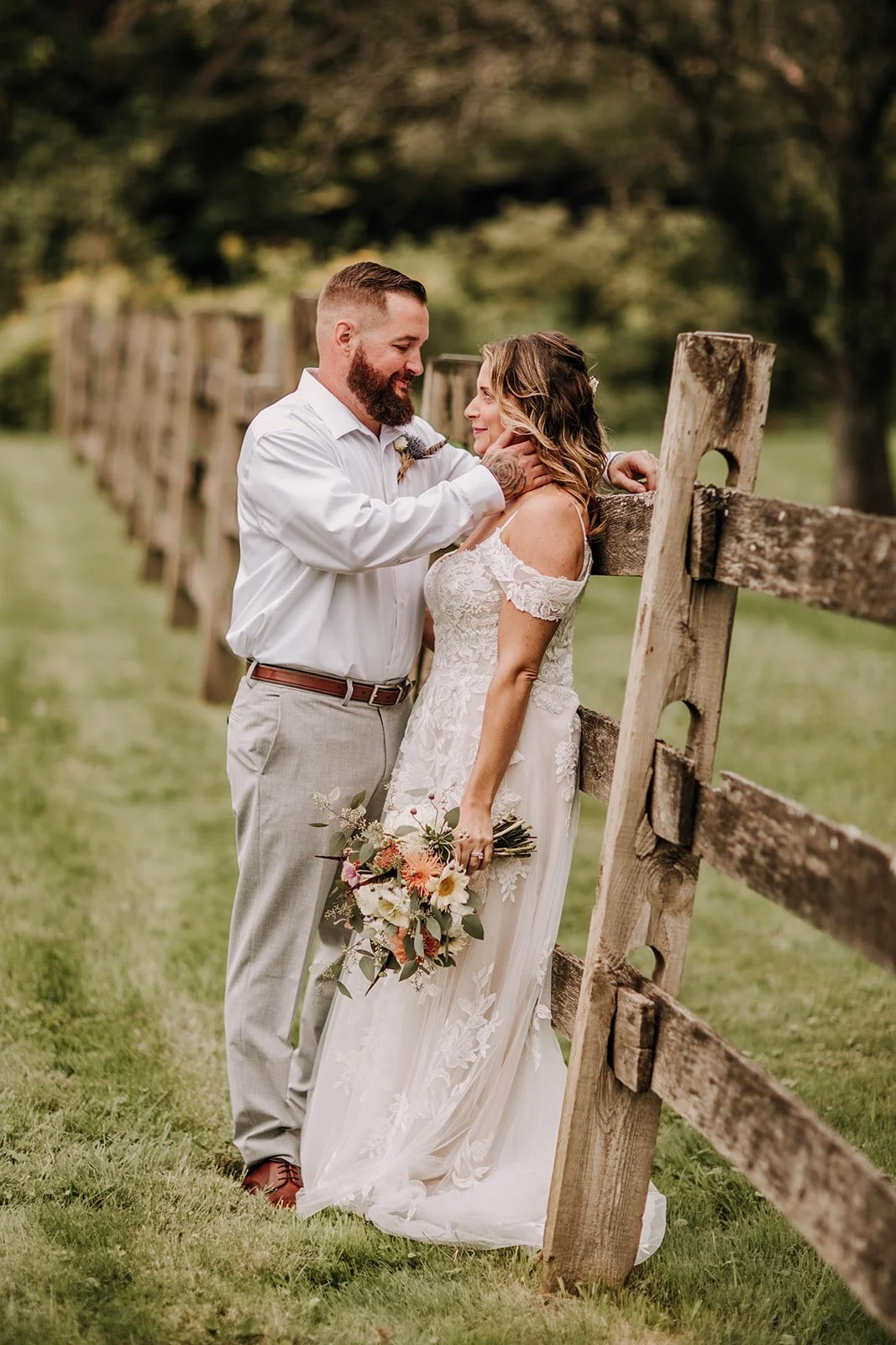 A bride and groom standing close together outdoors by a wooden fence, with the groom gently touching the bride's face, both smiling. The bride is holding a bouquet of flowers, wearing a lace wedding dress, and the groom is dressed in light-colored trousers and a white shirt. The background features green grass and trees.
