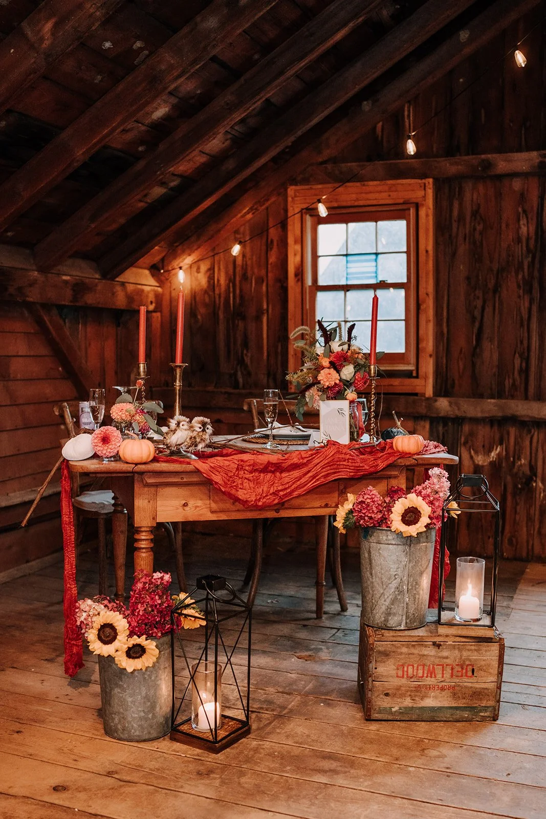 Rustic wooden table decorated with pink and orange flowers in metal buckets, tall pink candles, small pumpkins, and owl figurines, set in a cozy wooden room with string lights, a window, and lanterns with candles.