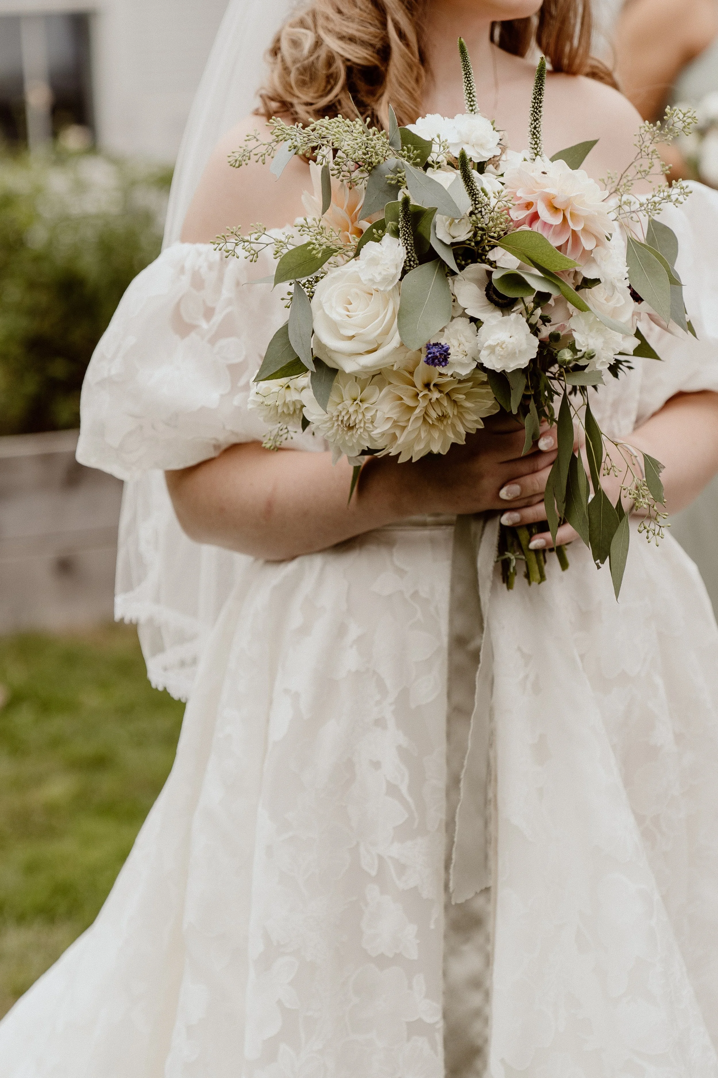 A person dressed in a white floral lace dress holds a bouquet of white and blush pink flowers with green foliage at a wedding or special event.