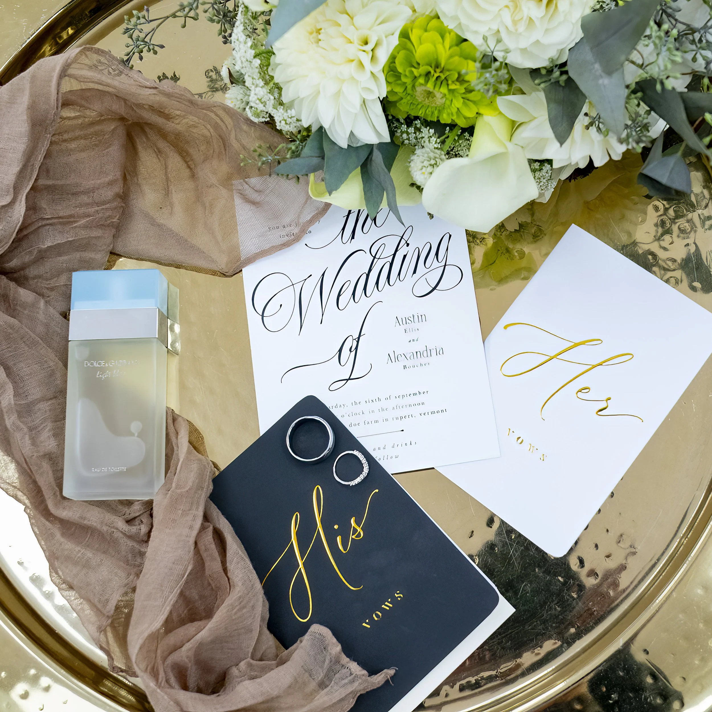 Wedding invitation suite with floral arrangement, two rings, and a small box on a reflective gold table. The items include a white wedding invitation with elegant black script, a white card with gold lettering, and a navy card with gold script, surrounded by beige fabric and a bouquet of white and green flowers.
