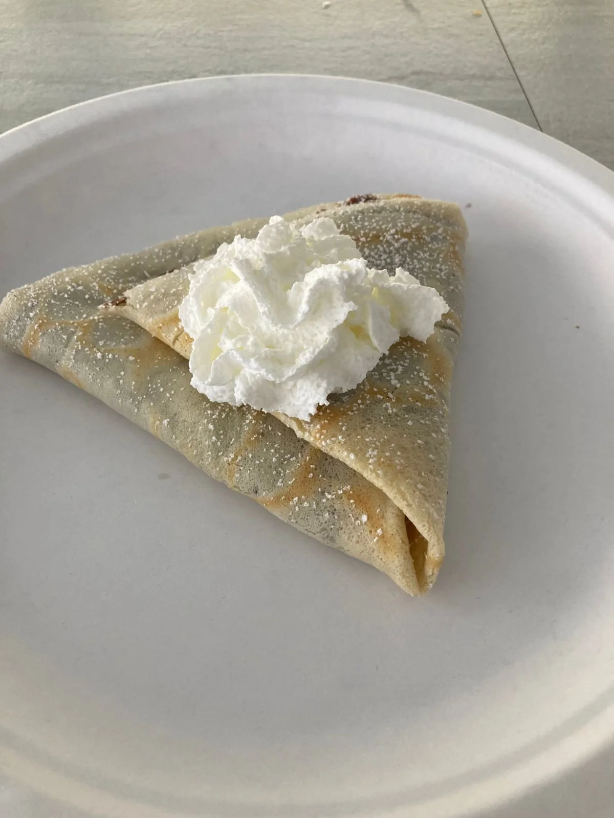 Crepe topped with whipped cream and powdered sugar on a white disposable plate.