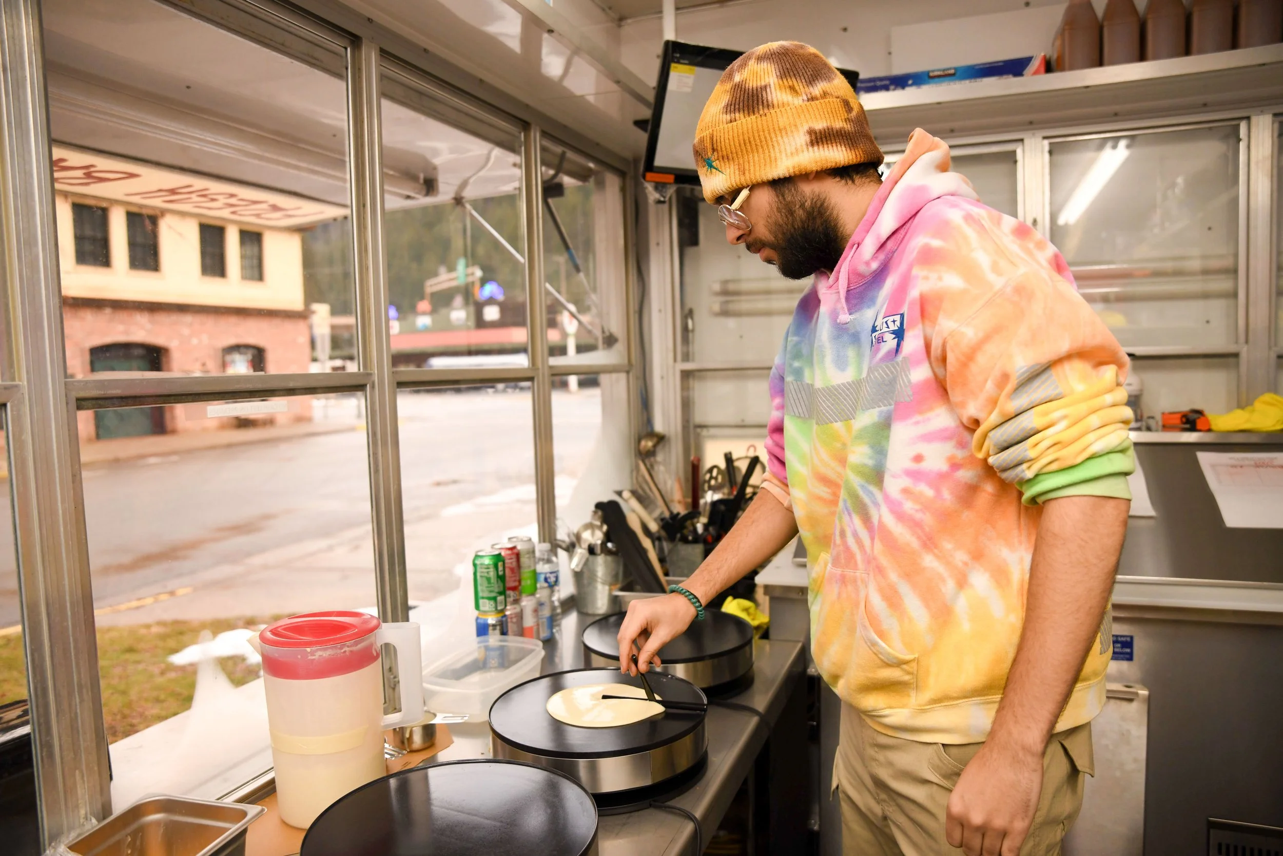 A man in a tie-dye hoodie, tan pants, glasses, and a yellow tie-dye beanie is cooking on a griddle at a food stand. The stand has various cans and kitchen tools on the counter, and there's a window looking out to a street with buildings.