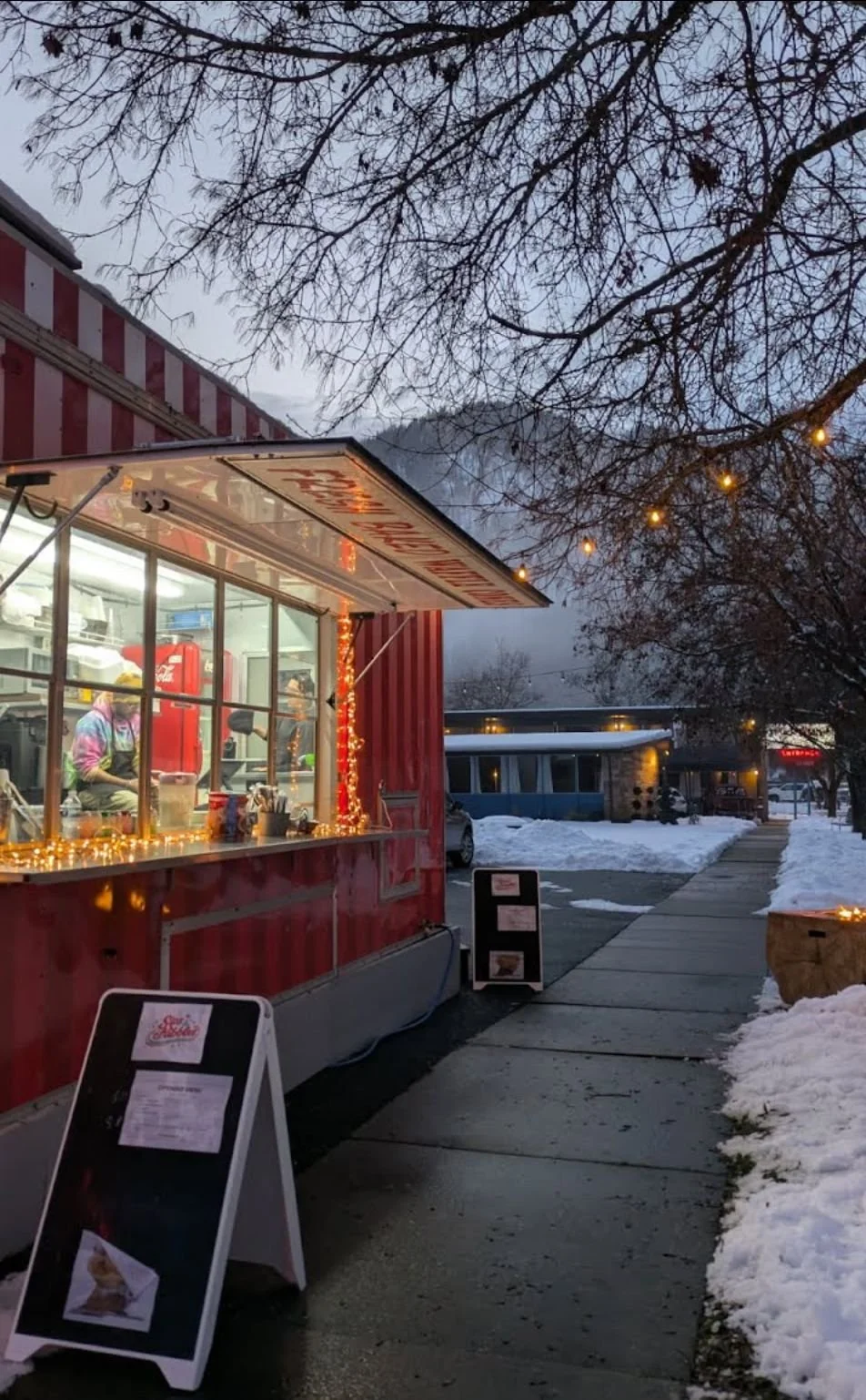 A snowy street scene at dusk featuring a red food stand decorated with string lights, with a worker inside preparing food, and a sidewalk with signs, trees, parked cars, and buildings in the background.